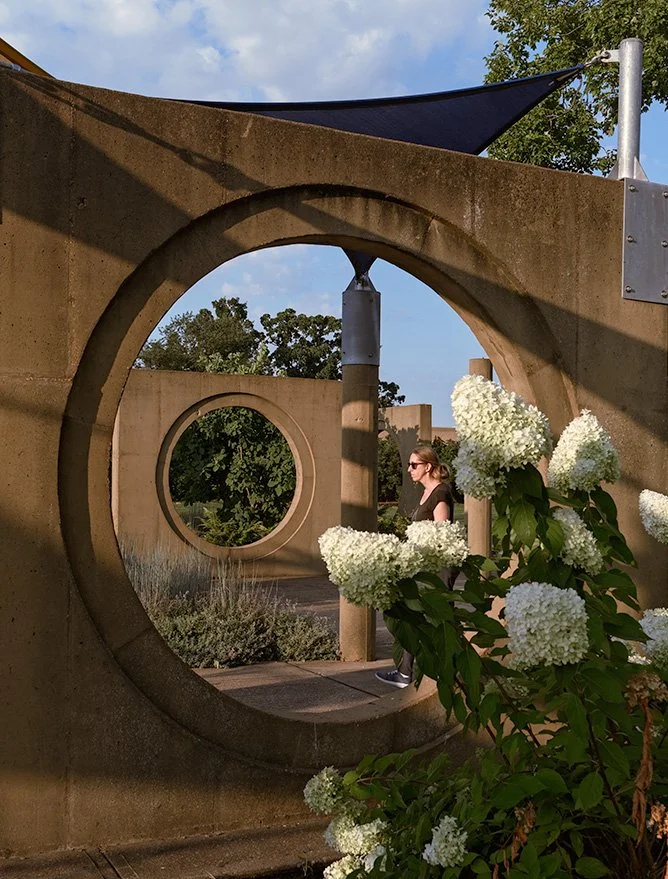 A woman walking outdoors, viewed through circular concrete openings in the wall, with white hydrangea flowers in foreground and trees in the background on a sunny day.