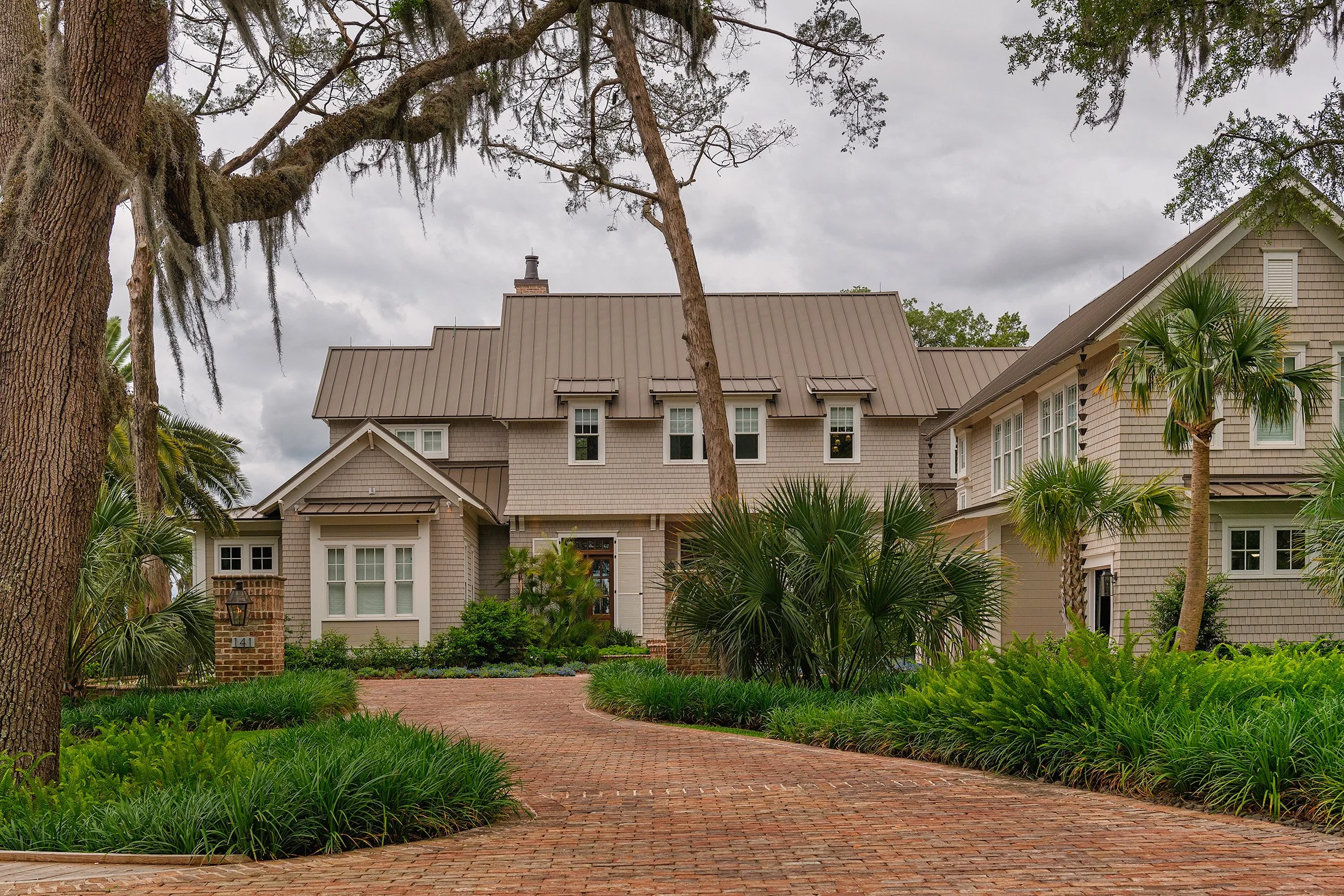 A large, multi-story house with beige siding, a metal roof, and multiple windows, surrounded by lush landscaping, palm trees, and a brick driveway on a cloudy day.