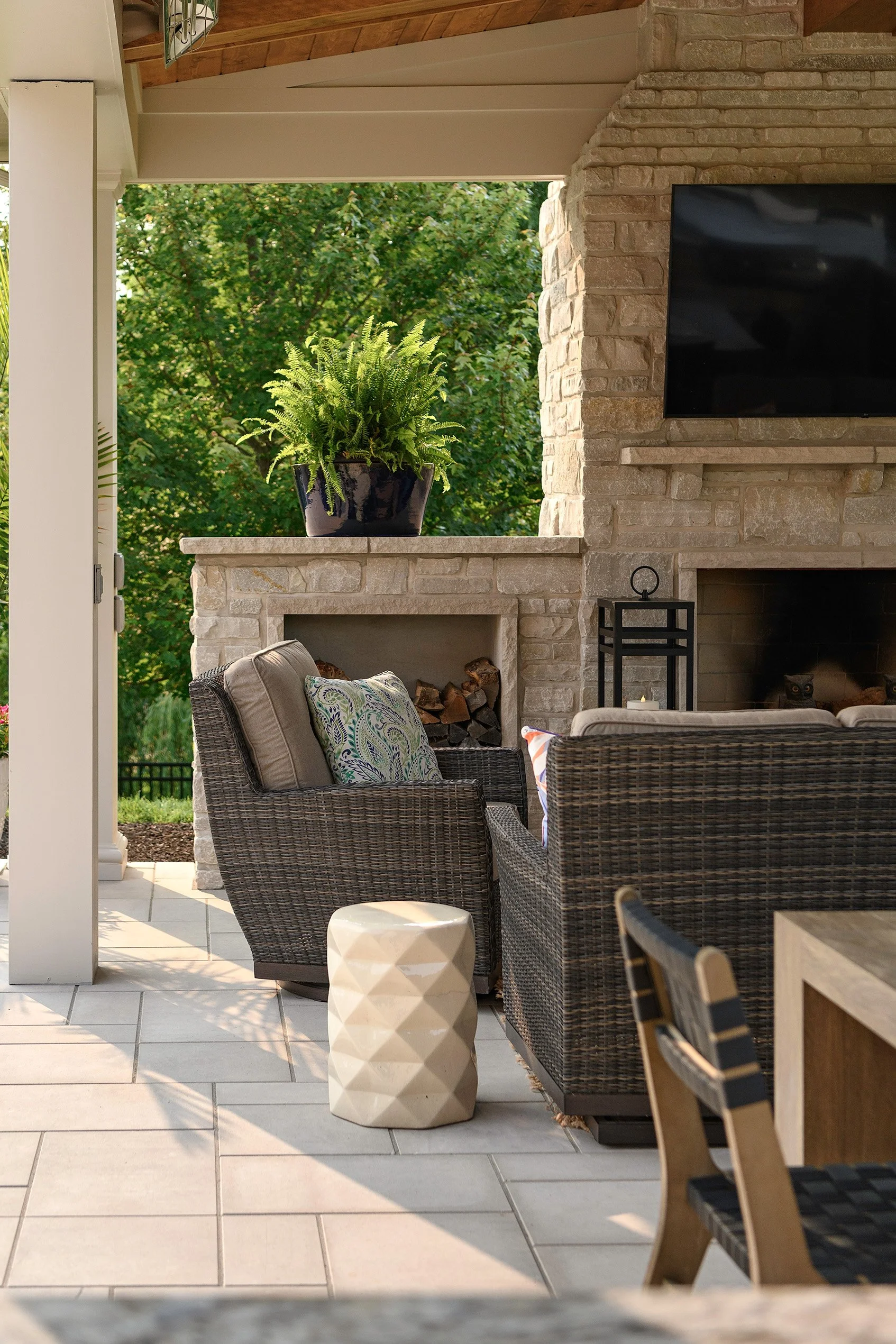 Outdoor patio with wicker chairs, beige cushions, a ceramic side table, a stone fireplace with logs, a flat-screen TV, a potted fern, and trees in the background.