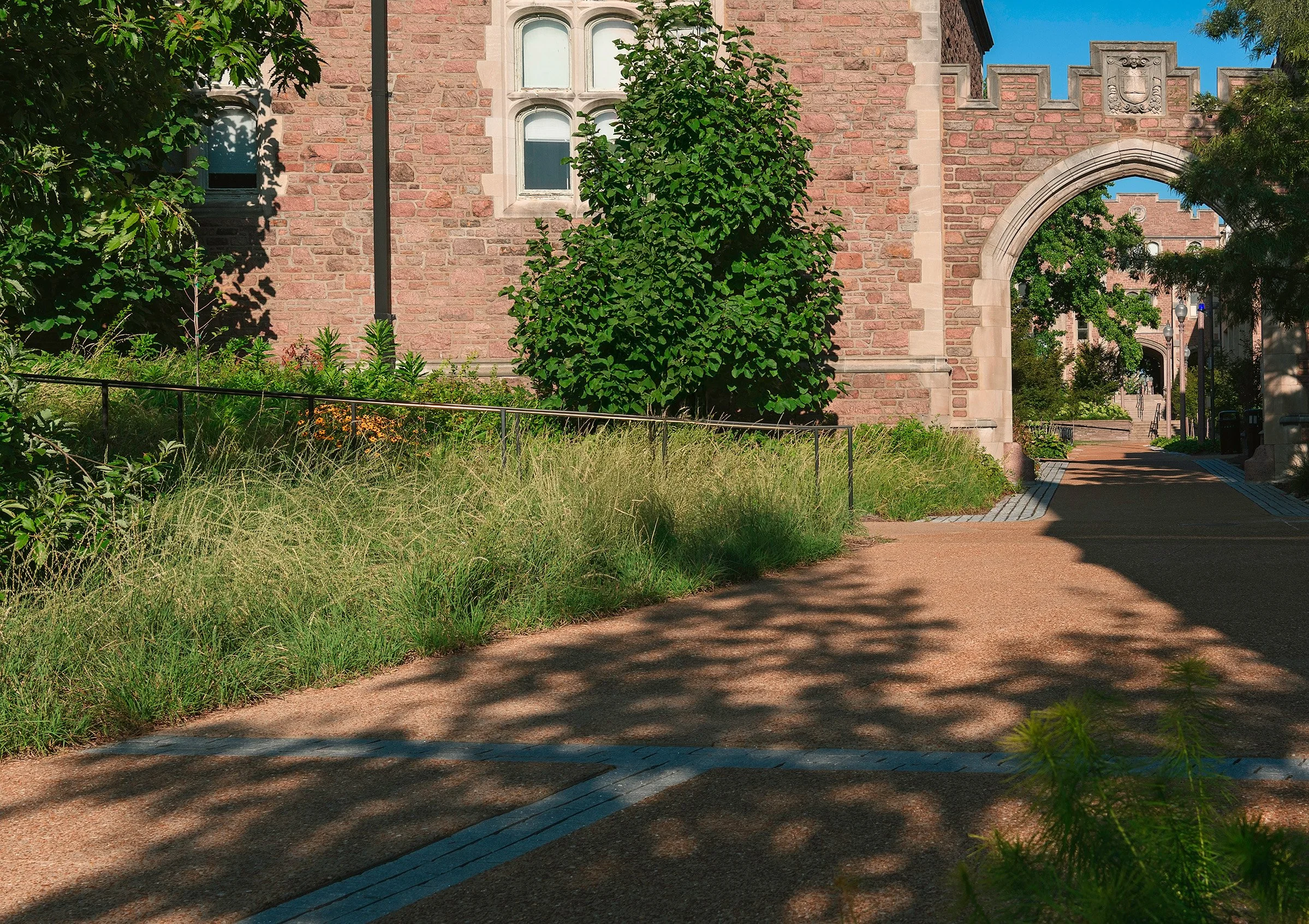 A stone building with arched doorways and windows, surrounded by greenery including trees and grass, with a paved pathway leading under an archway.