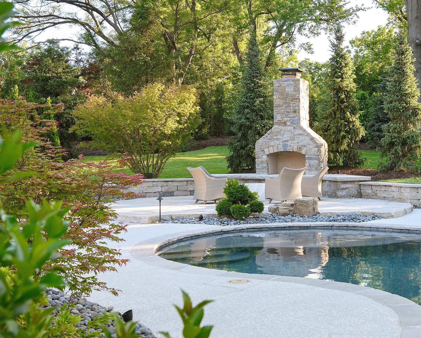 A backyard patio with a fire pit and beige chairs, located near a swimming pool, with lush green trees and shrubs in the background.