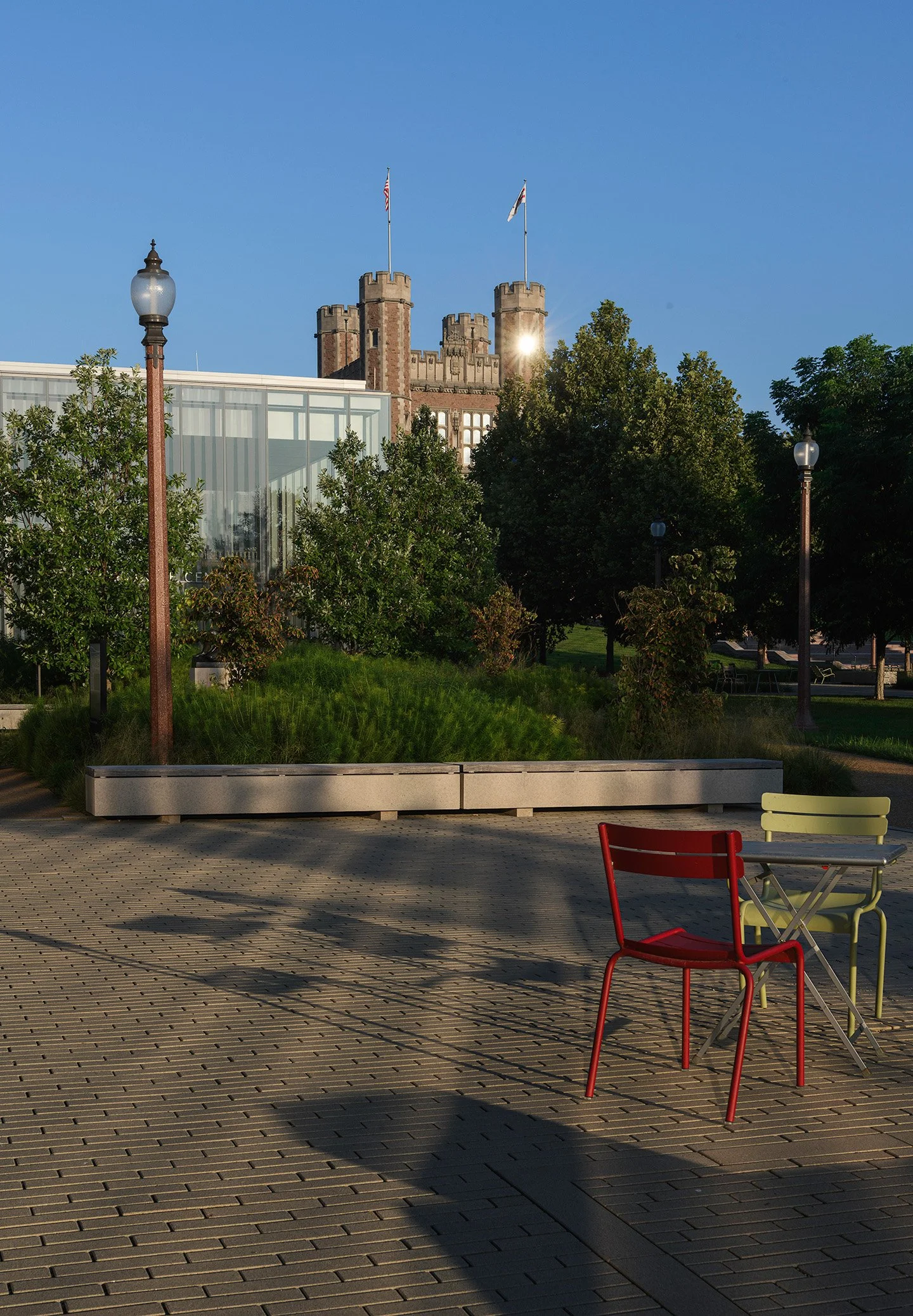 Urban park with colorful chairs, trees, and shadows, with a castle in the background under a clear blue sky.