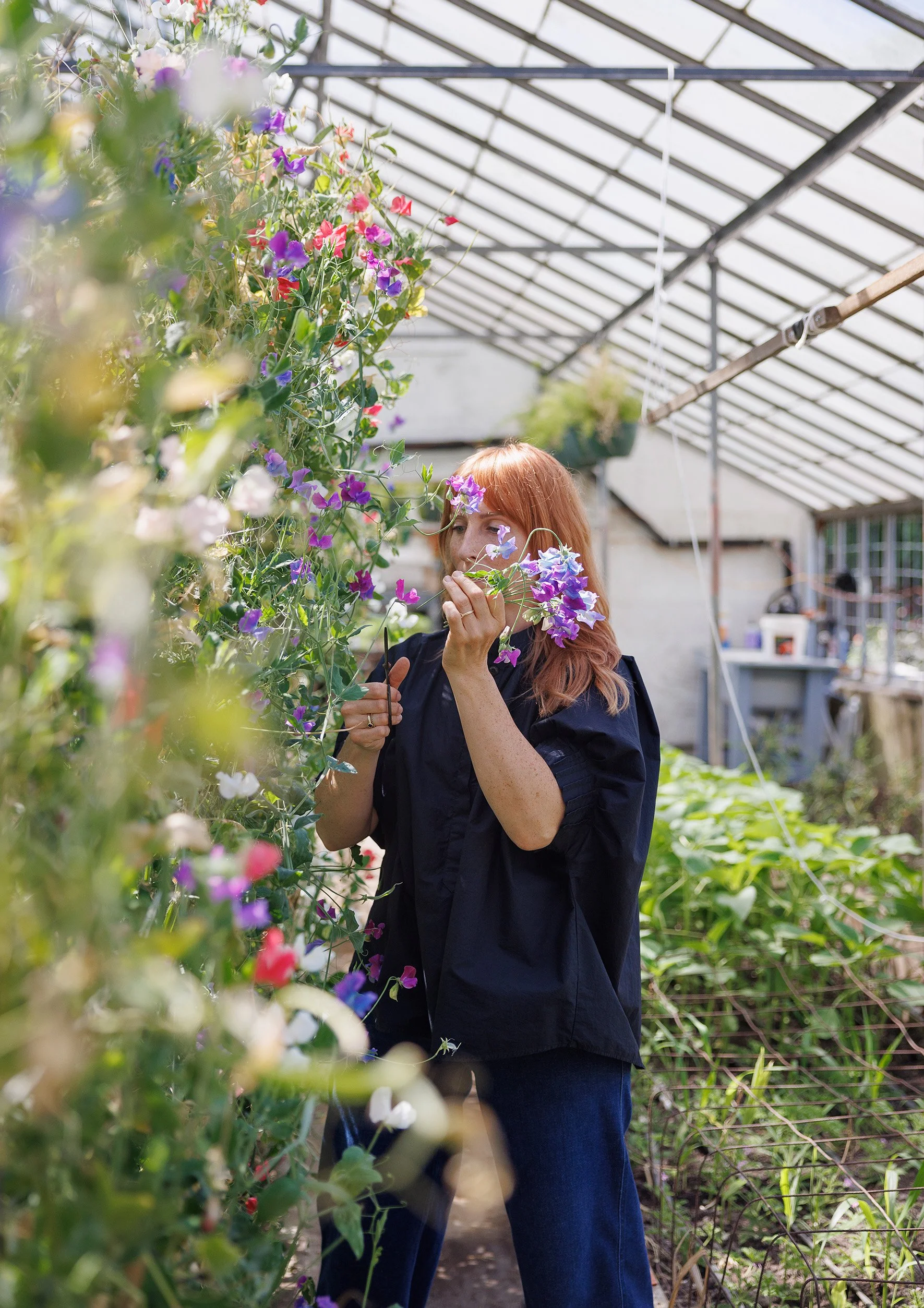 A woman with red hair wearing a black jacket and jeans smelling purple and pink flowers in a greenhouse.