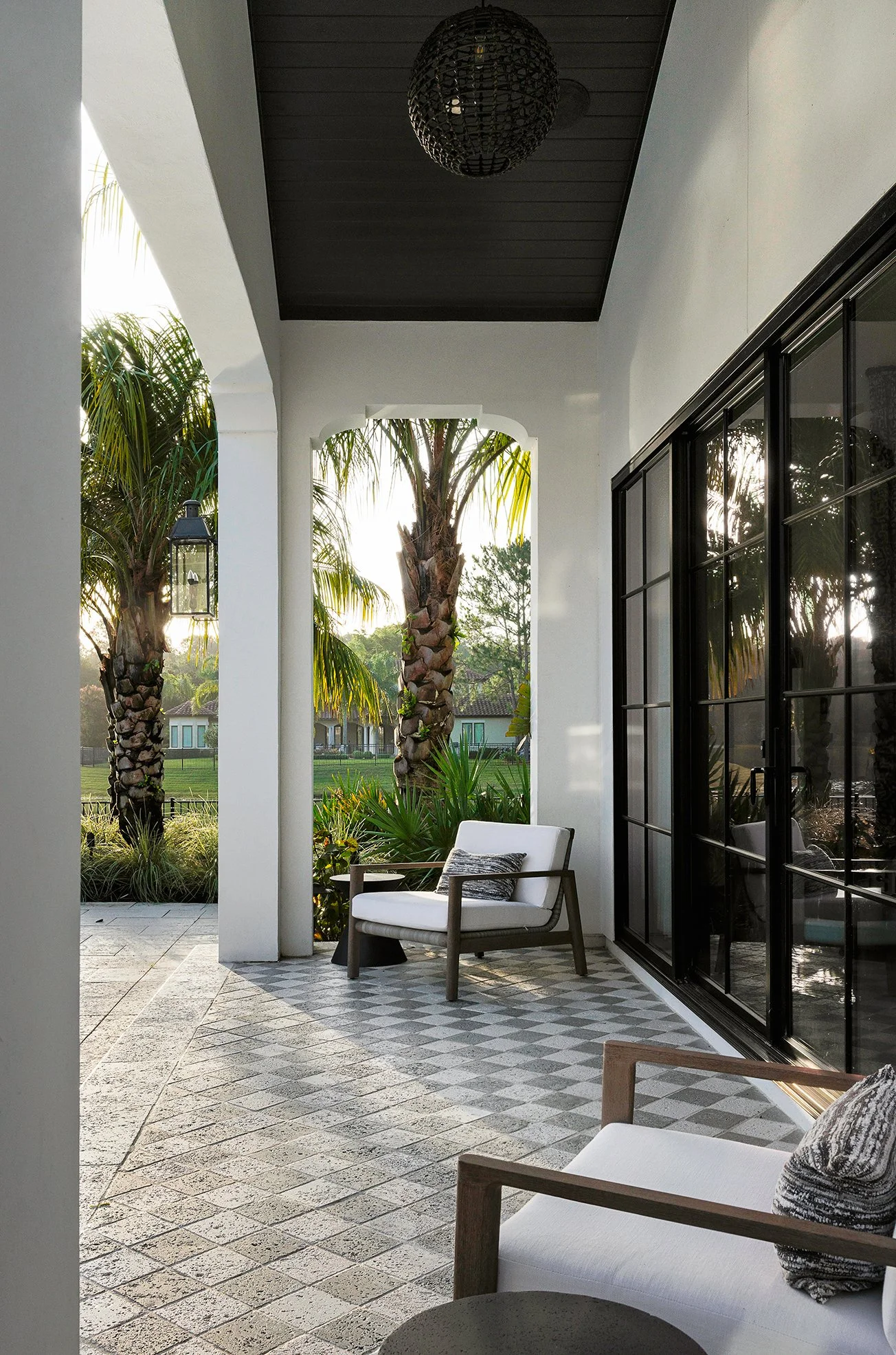 Patio with white chairs, decorative pillows, large windows, and palm trees outside.