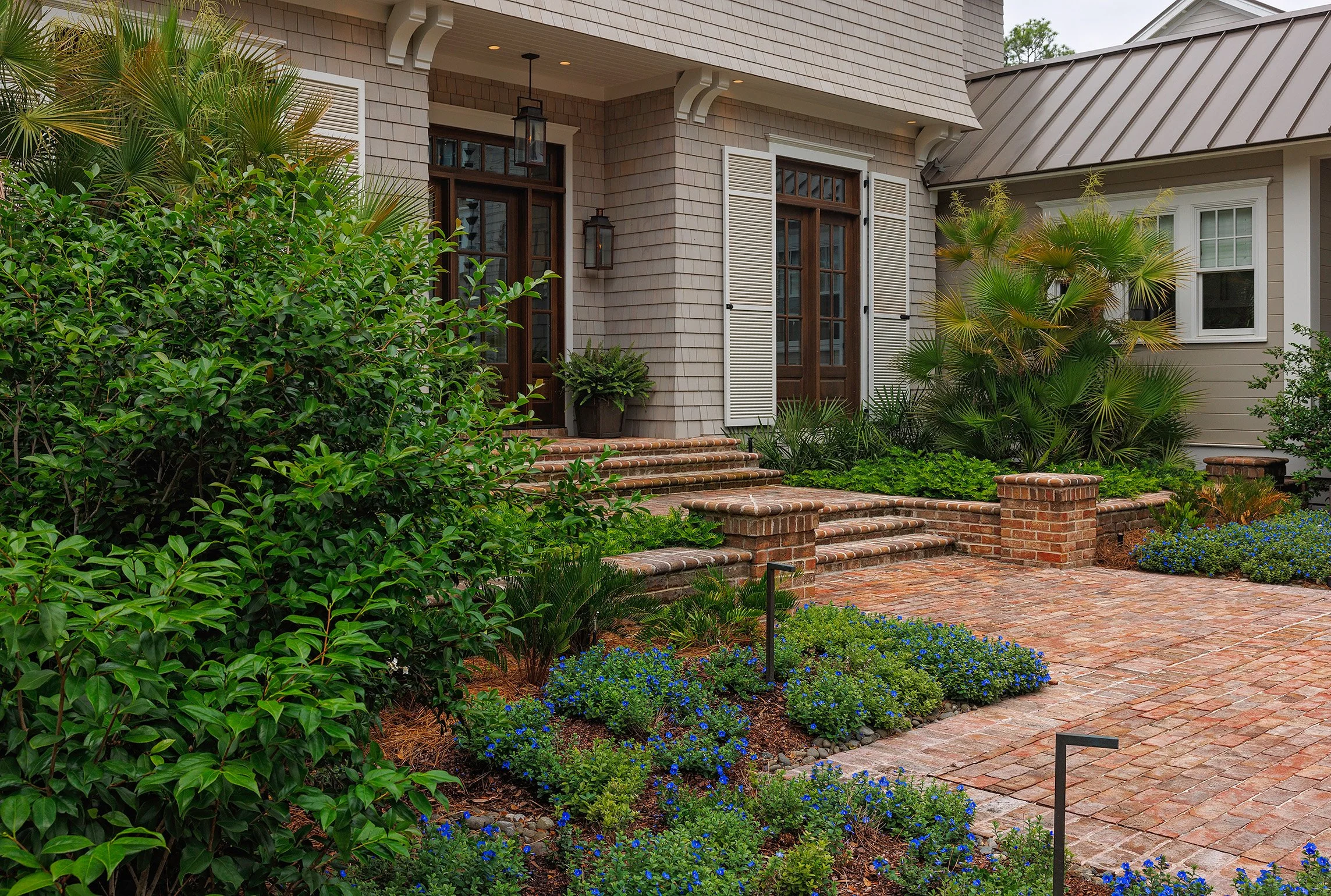 Front entrance of a house with brick steps, a brick patio, and landscaped garden with shrubs and plants.