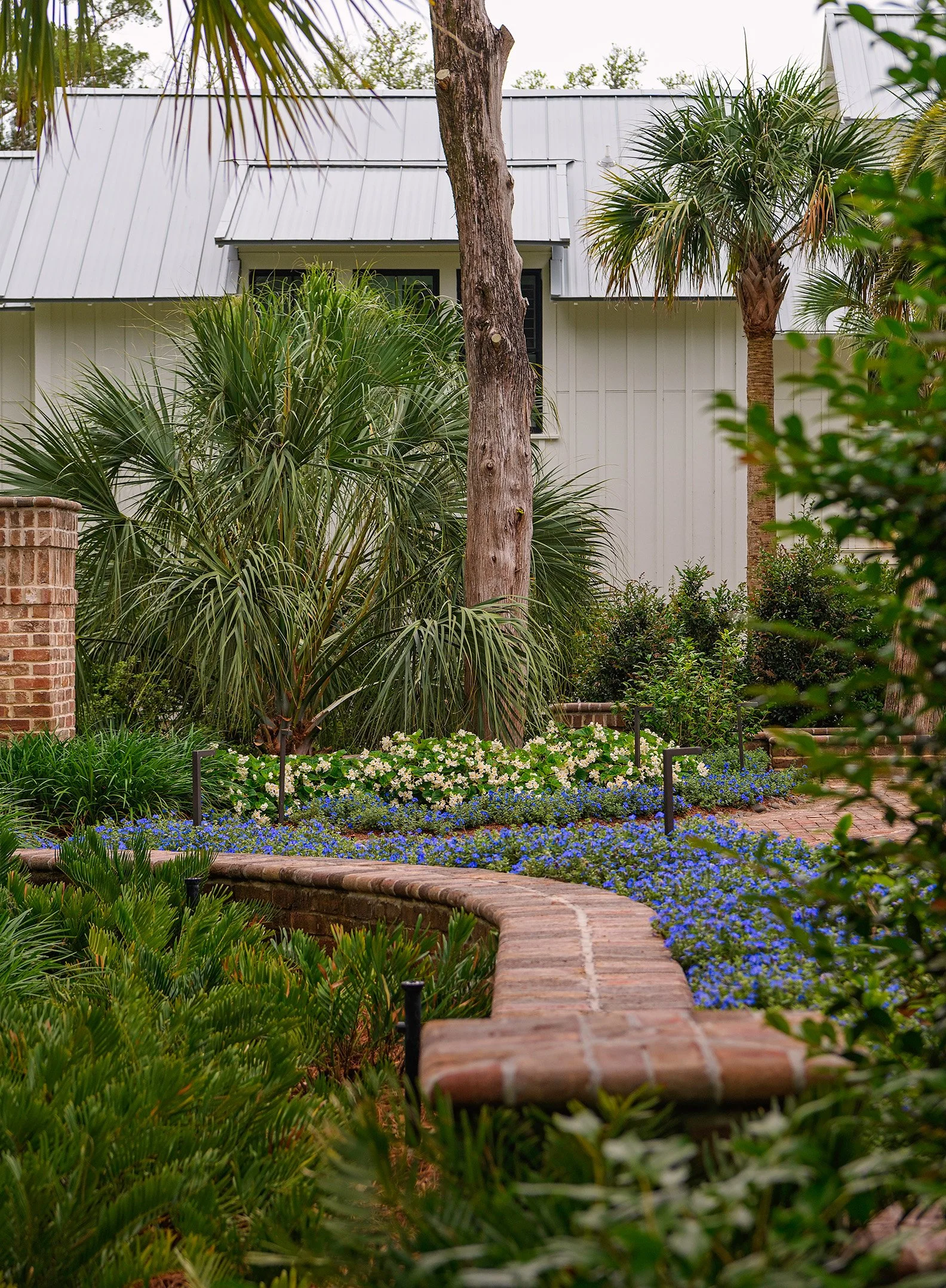 A garden with lush green plants, bushes, and flowers, along with a brick pathway and brick border wall. There are trees and a house with a metal roof in the background.