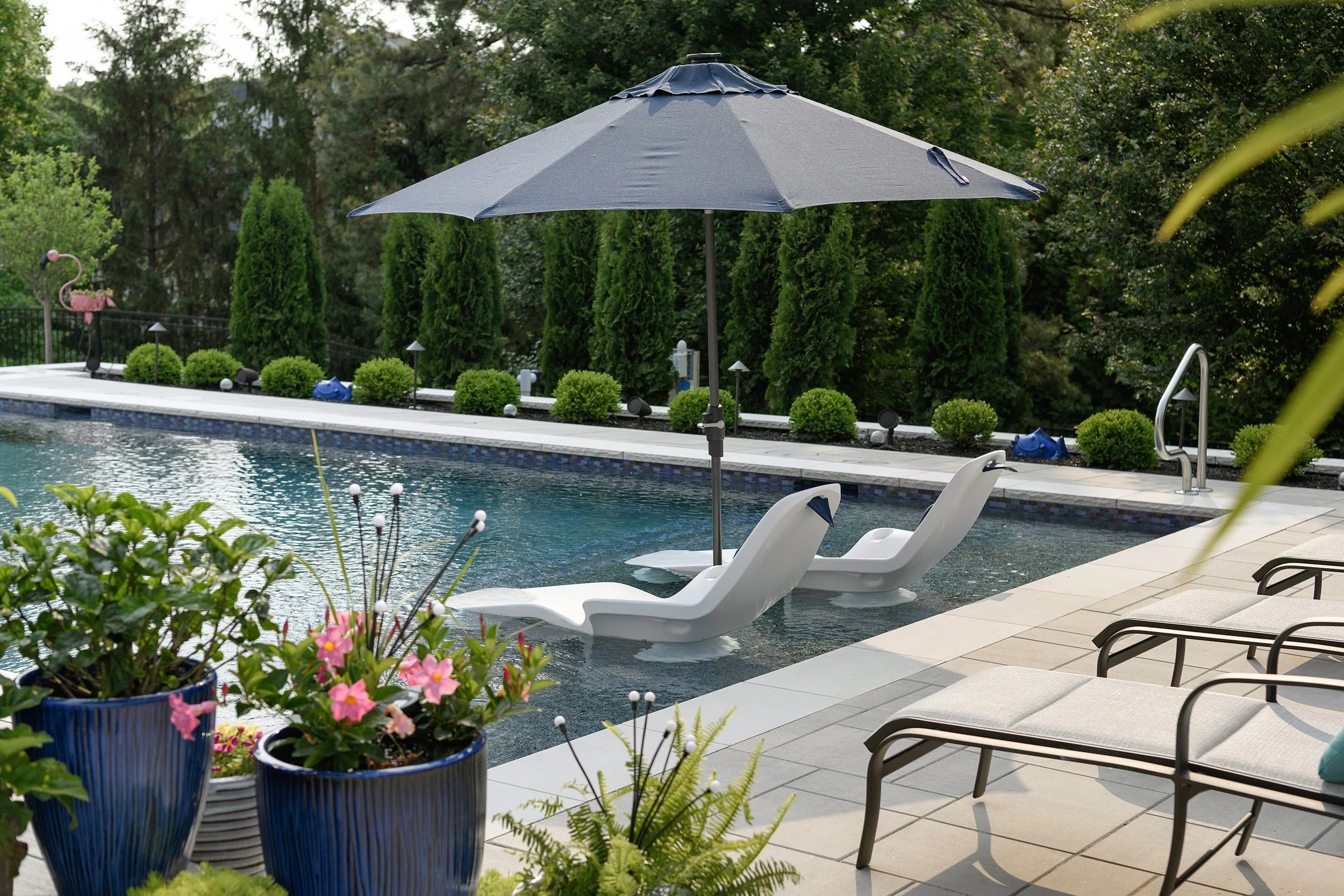 Swimming pool with two white lounge chairs under a large gray umbrella, surrounded by potted plants, trees, and outdoor furniture.