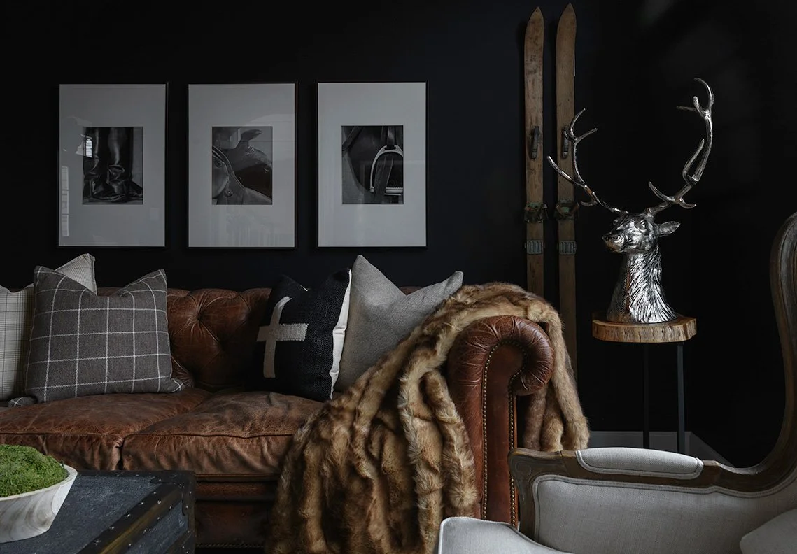 Interior design photography of a living room with a brown leather sofa, decorative pillows, a faux fur throw, black wall with three framed black-and-white photos, vintage skis, and a deer head sculpture with antlers on a stand.