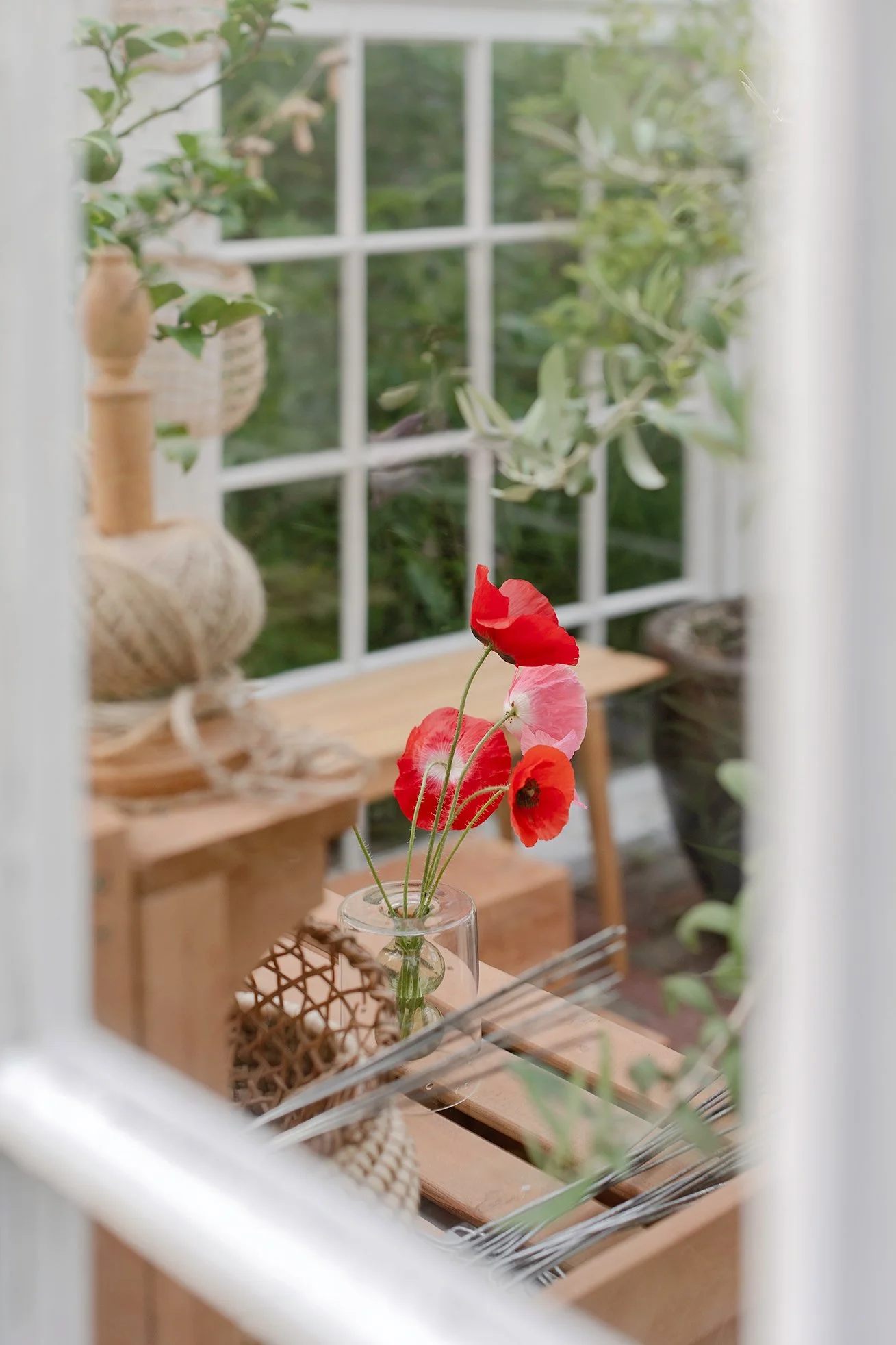 Scene of a windowsill with a small glass vase holding red and pink poppy flowers, surrounded by wicker and wooden decorative items, with a view of green trees outside through the window.