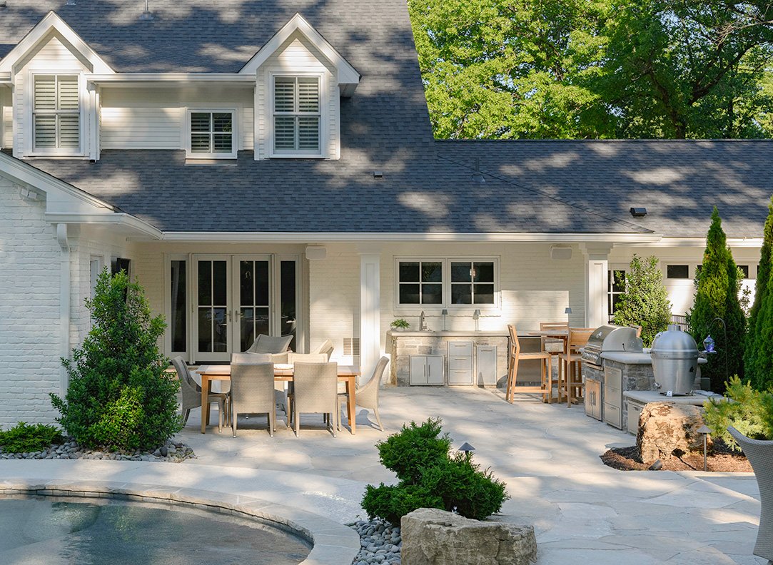 Backyard patio with outdoor dining table, chairs, outdoor kitchen with grill, and landscaping with bushes and trees.