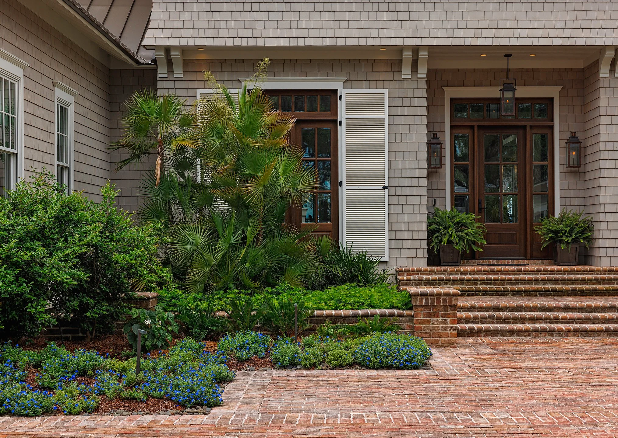 Front entrance of a house with brick steps, wooden door, potted plants, and landscaped garden with green plants and blue flowers.