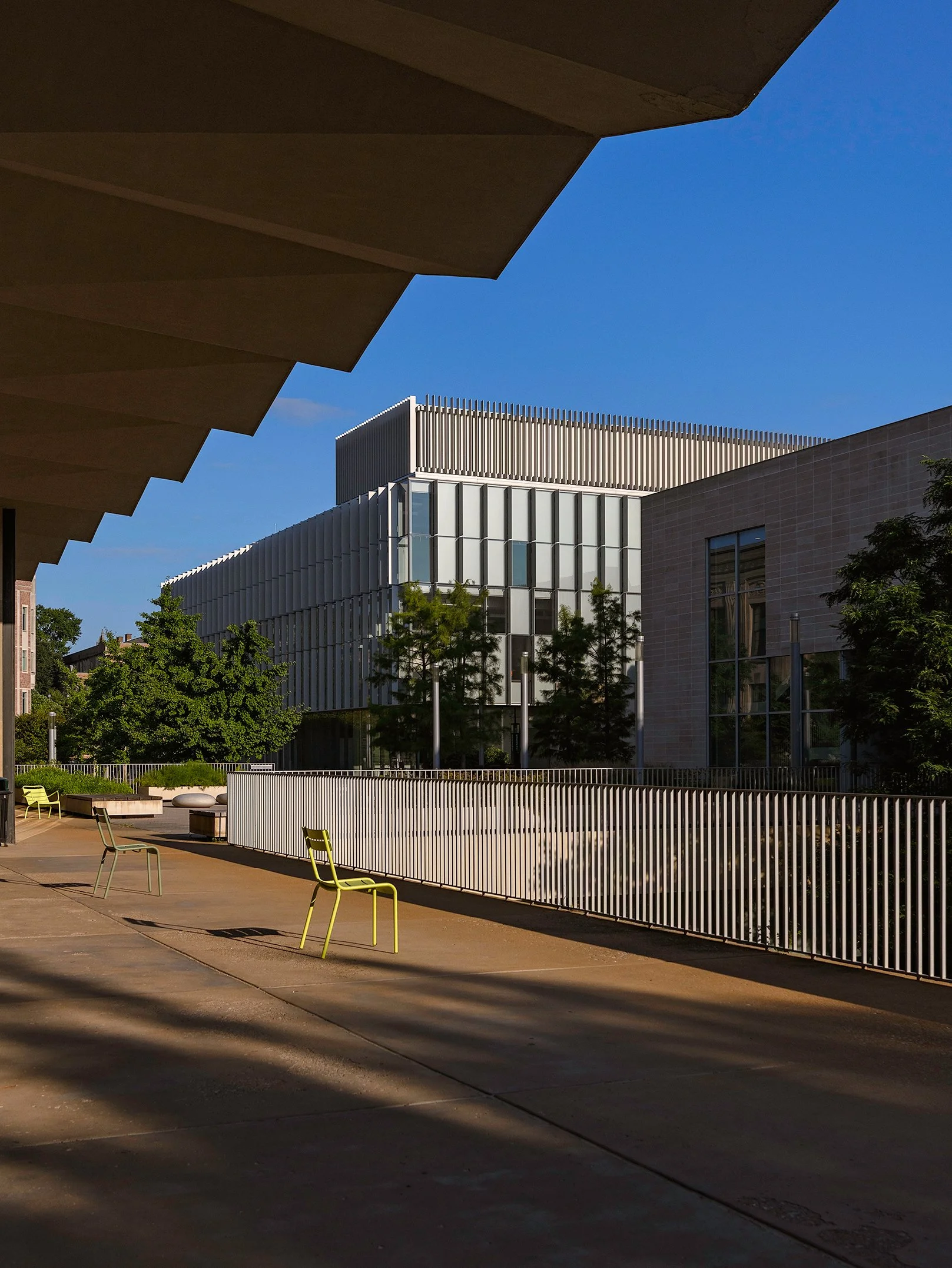 Modern building with vertical white slats and glass windows, trees, and seating area with colorful chairs, under a blue sky.