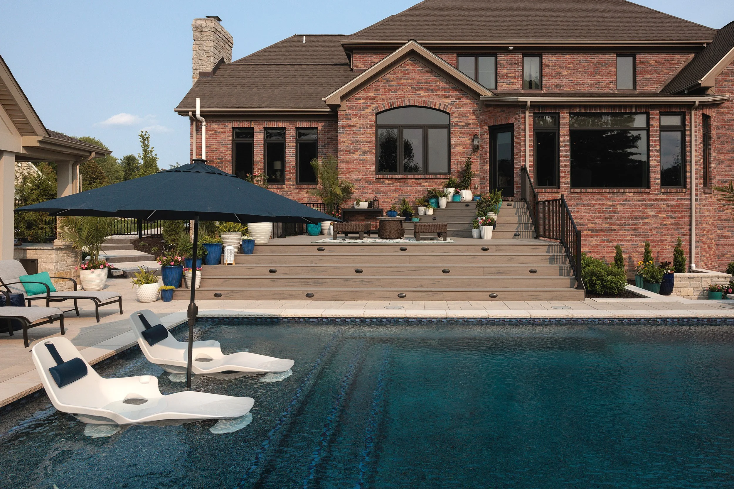 Backyard view of a brick house with a large deck and pool, surrounded by potted plants and outdoor furniture, including lounge chairs and umbrellas.