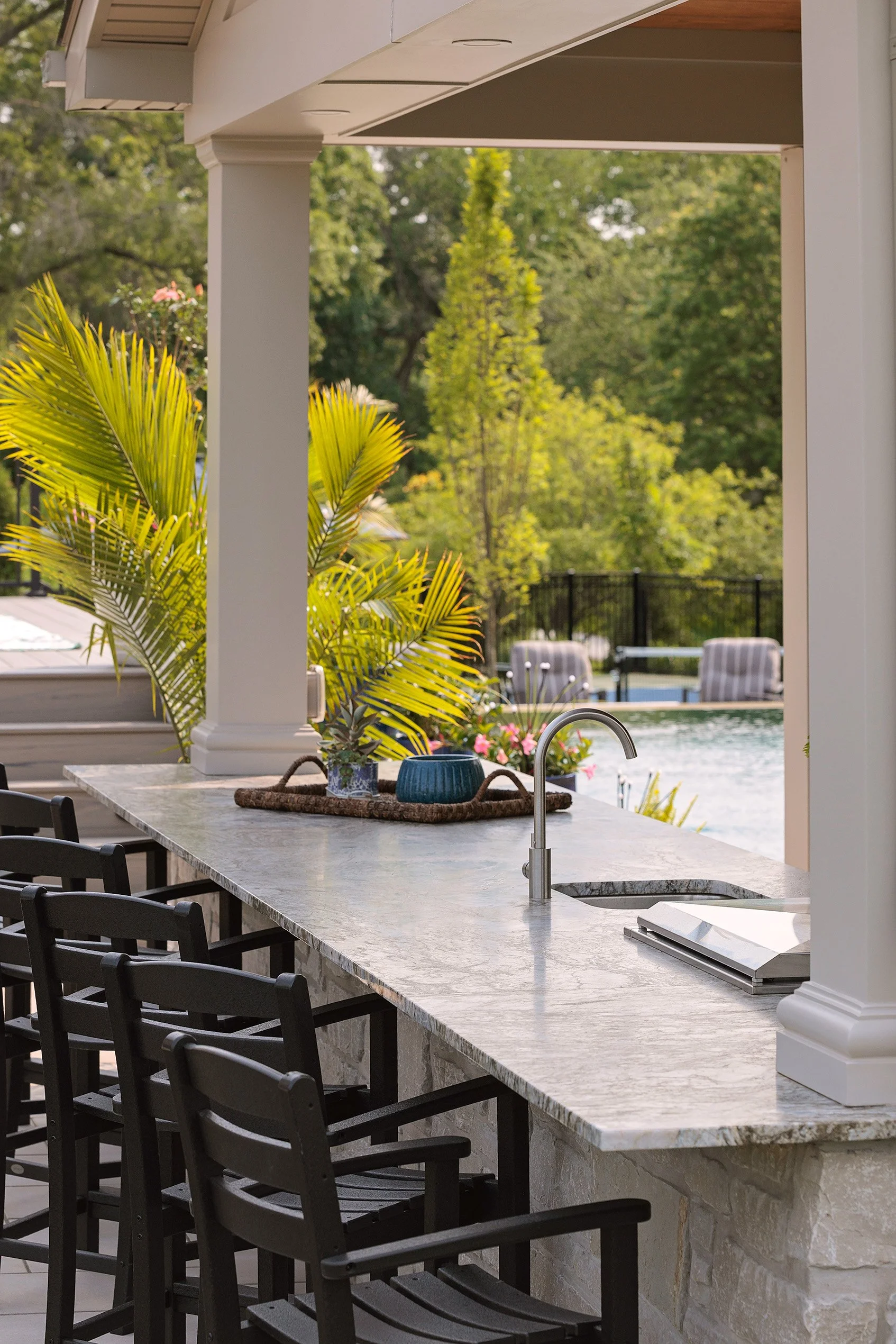 An outdoor kitchen with a marble countertop, black chairs, a sink, potted plants, and a pool in the background surrounded by trees.