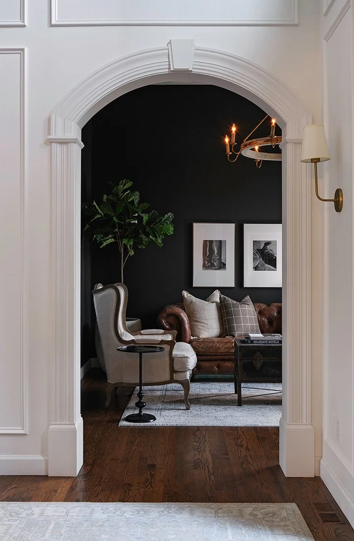 Interior design photography of a view of a cozy living room seen through an arched doorway, featuring a tan armchair, a brown leather sofa with pillows, a tall green plant, a black wall with framed artwork, and a chandelier.
