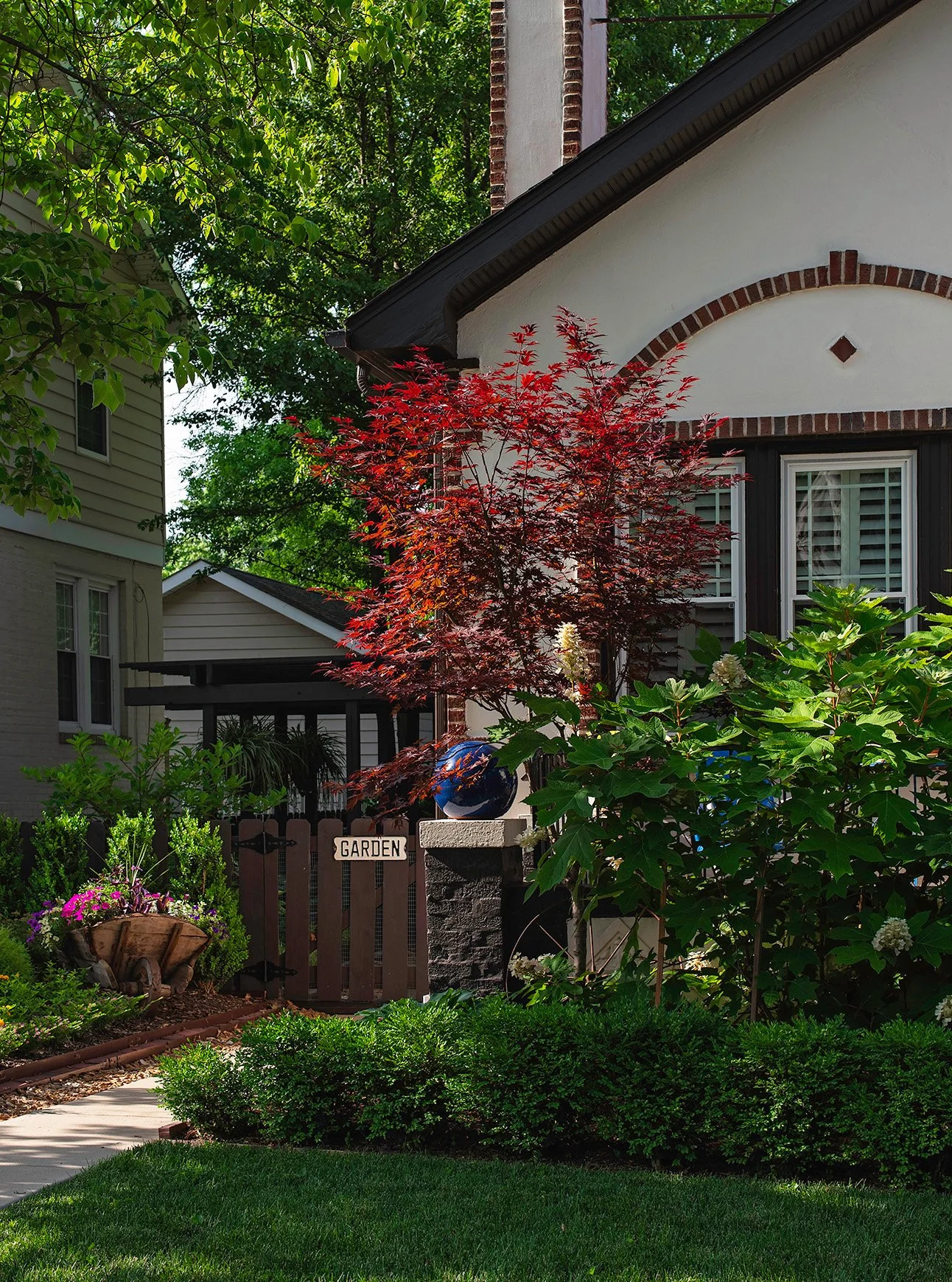 A front yard garden with a red-leafed tree, green bushes, and a flower bed, in front of a house with white walls and black trim, and a wooden gate marked "GARDEN."