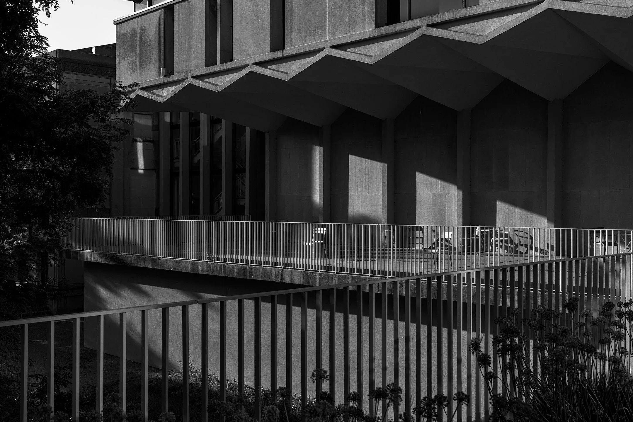 Black and white photograph of a modern building with large concrete walls and geometric shadows. There is a balcony with metal railings and empty chairs, and some plants in the foreground.