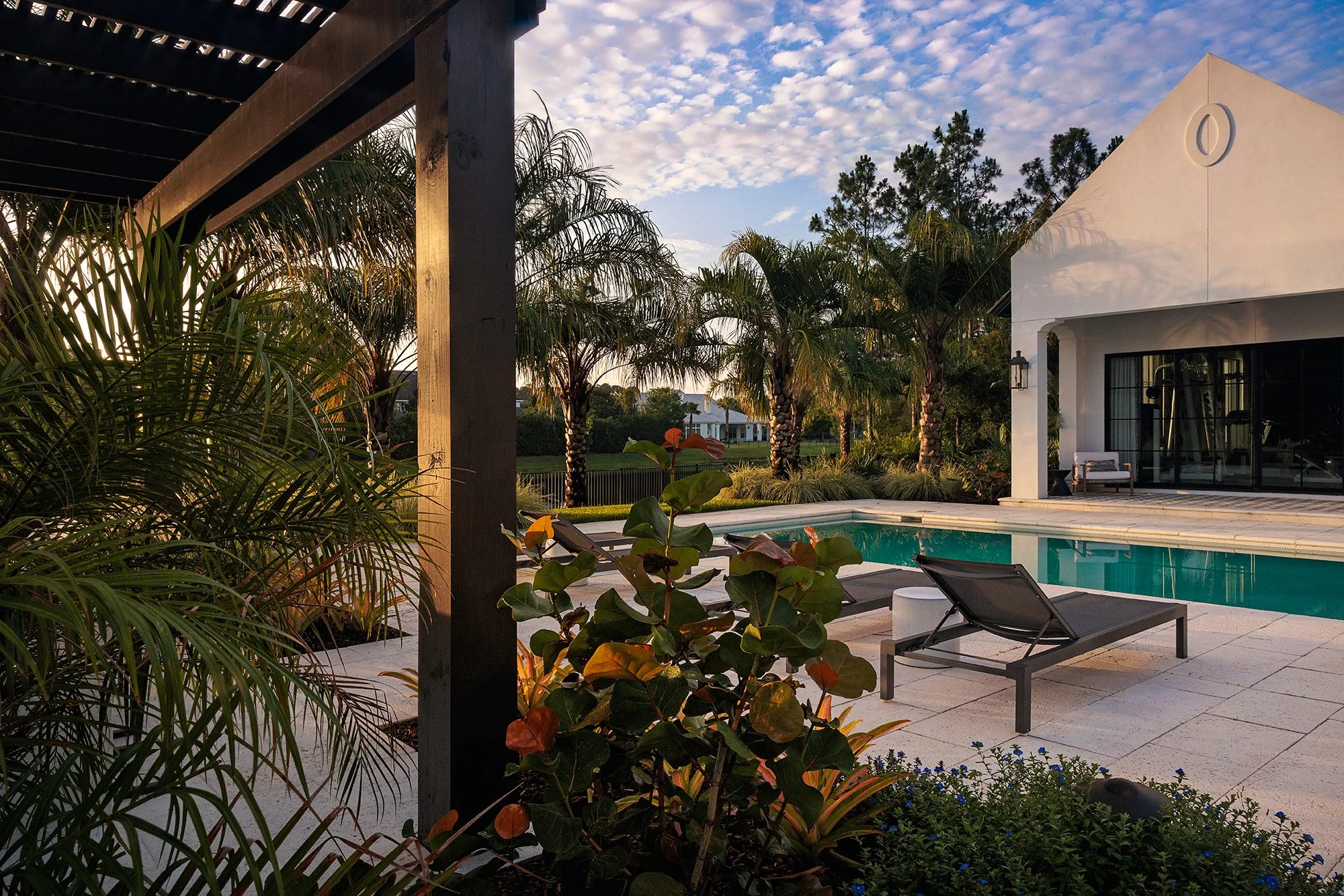 Luxury backyard with swimming pool, lounge chairs, surrounding palm trees, a modern white house, and a pergola structure at sunset.