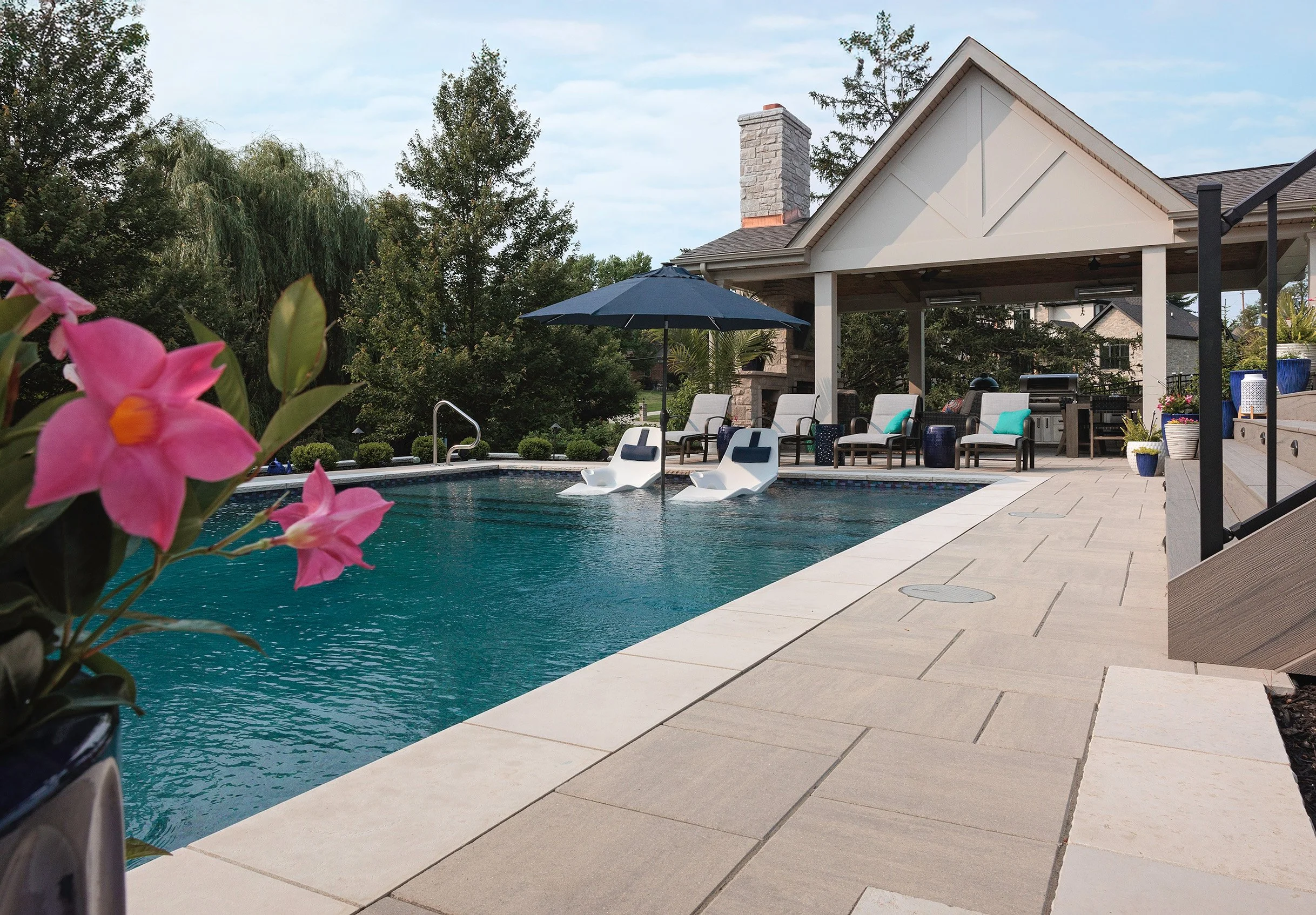 Backyard swimming pool with lounge chairs, umbrella, and a pavilion, surrounded by trees and a house in the background.