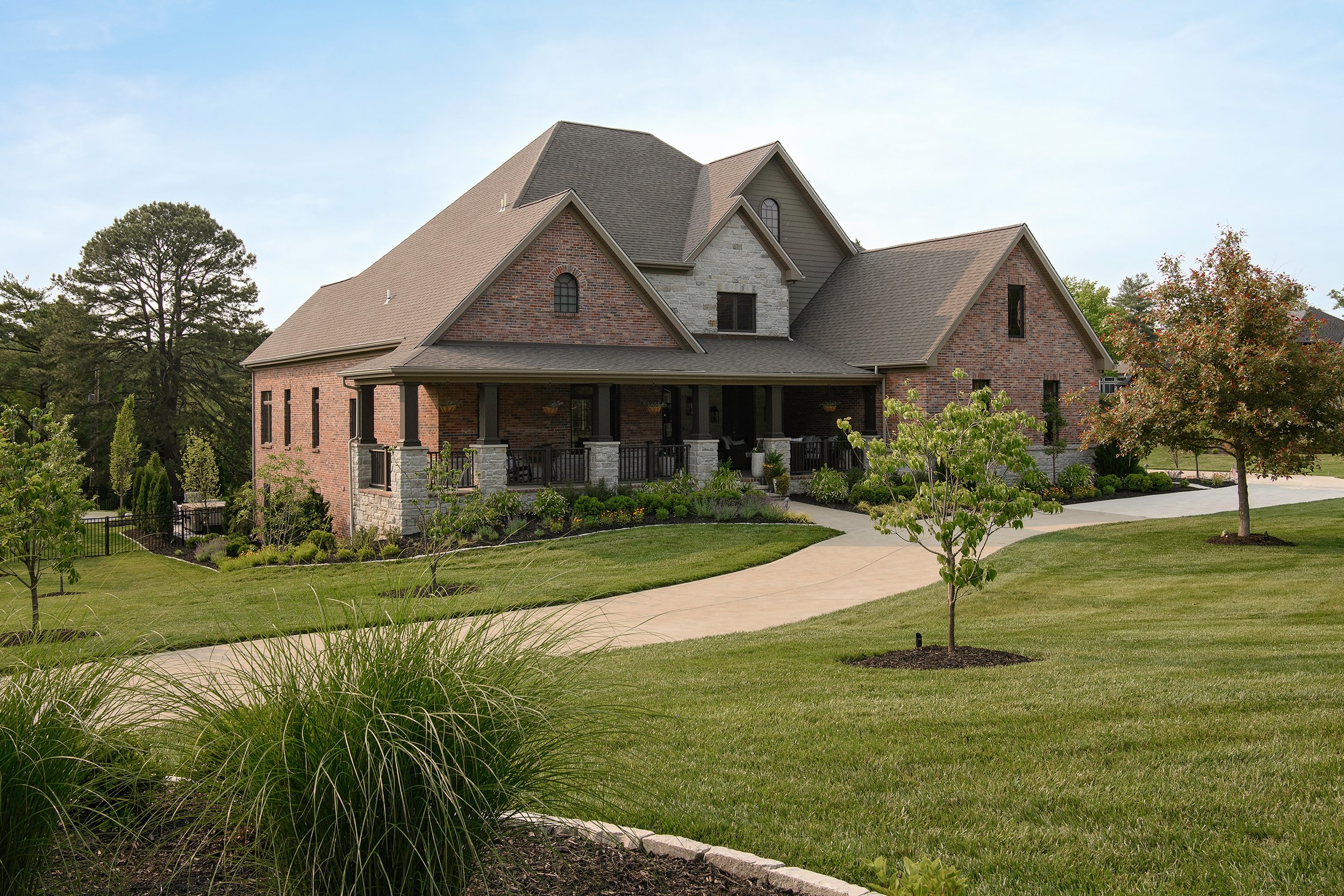A large brick house with a well-maintained lawn, trees, and a curved concrete driveway under a clear sky.