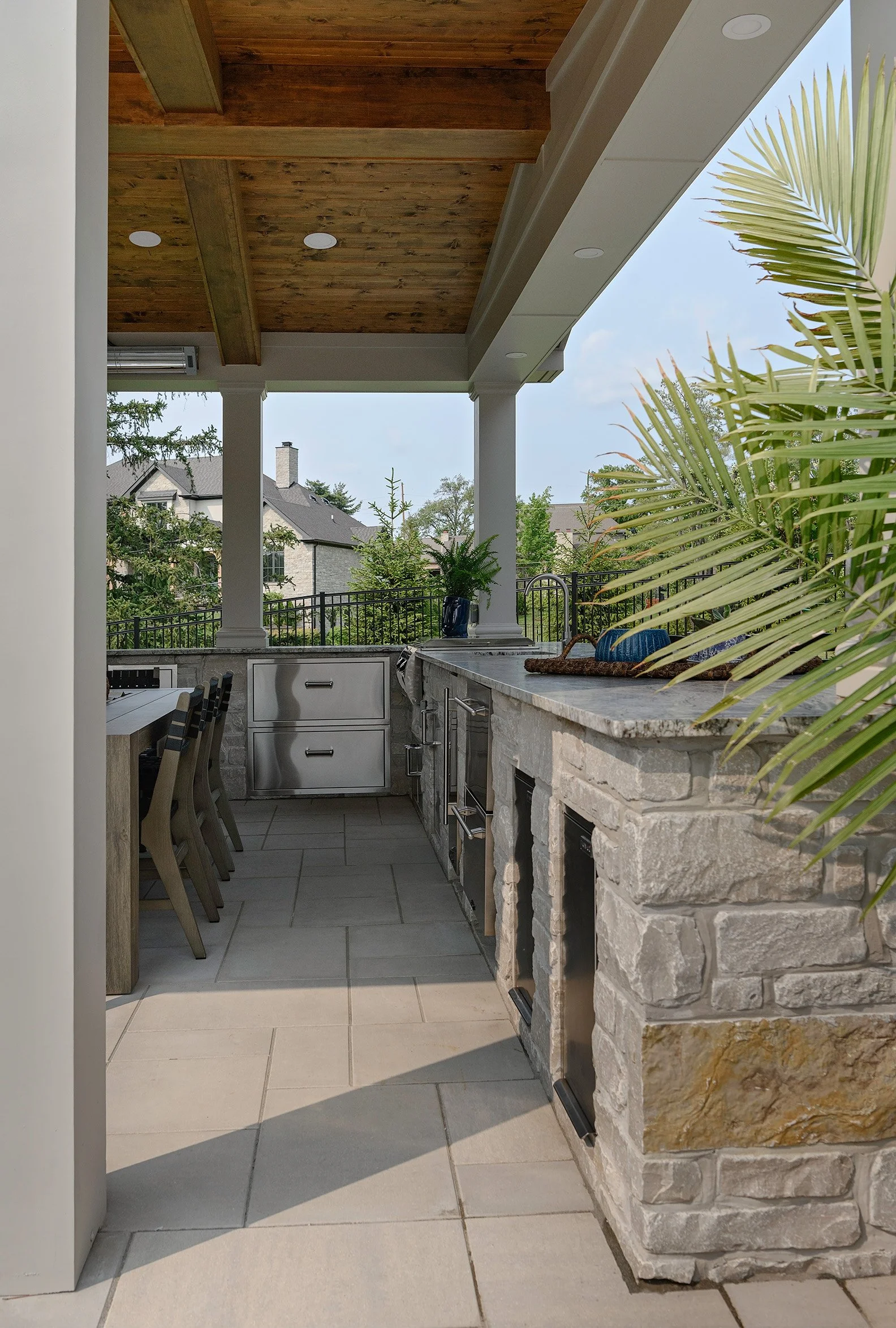 Outdoor kitchen with stone countertops, stainless steel appliances, and wooden ceiling beams on a covered patio.