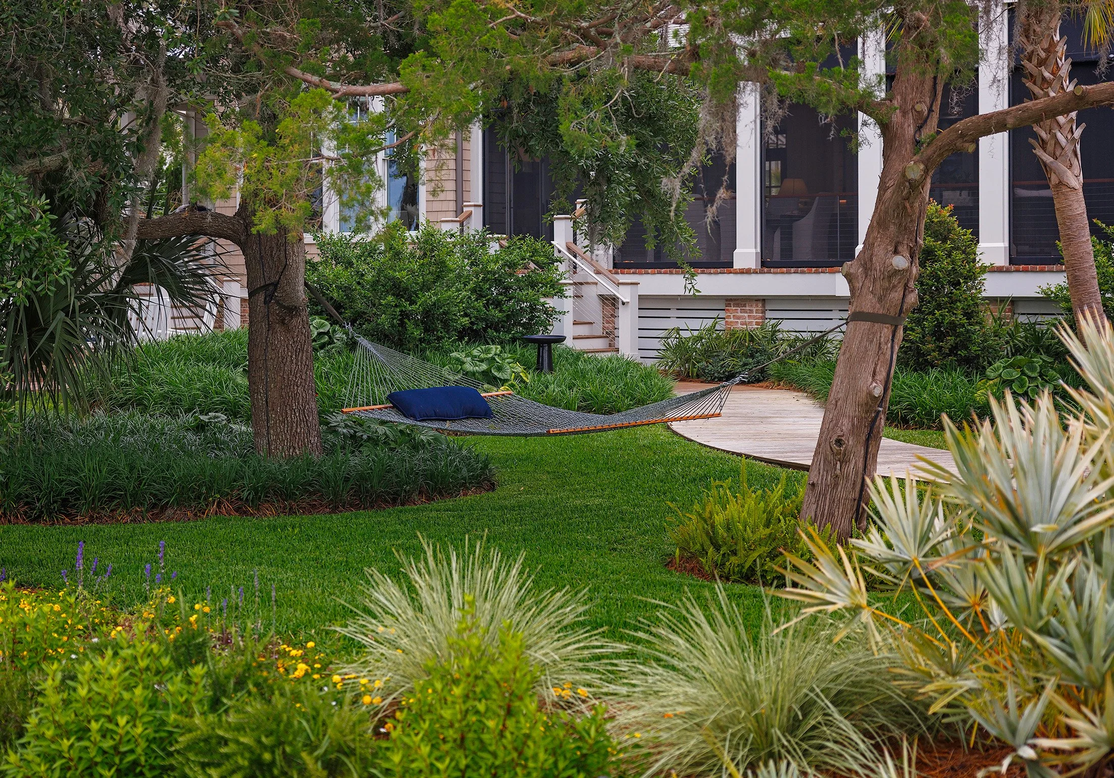 A backyard garden with lush green grass, various plants, and trees. A hammock with a pillow is tied between two trees. House with a porch and steps is visible in the background.