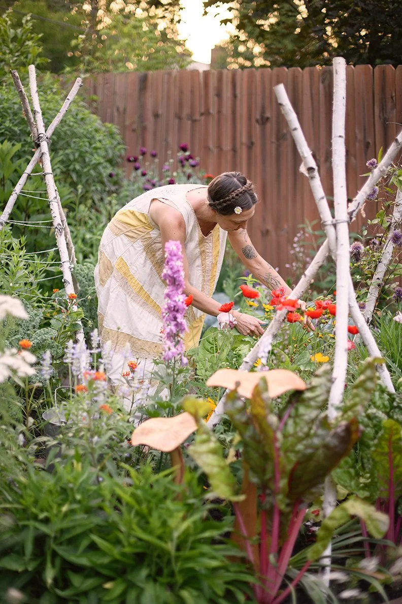 A woman with a flower in her hair tending to colorful flowers and plants in a garden, with a wooden fence in the background.