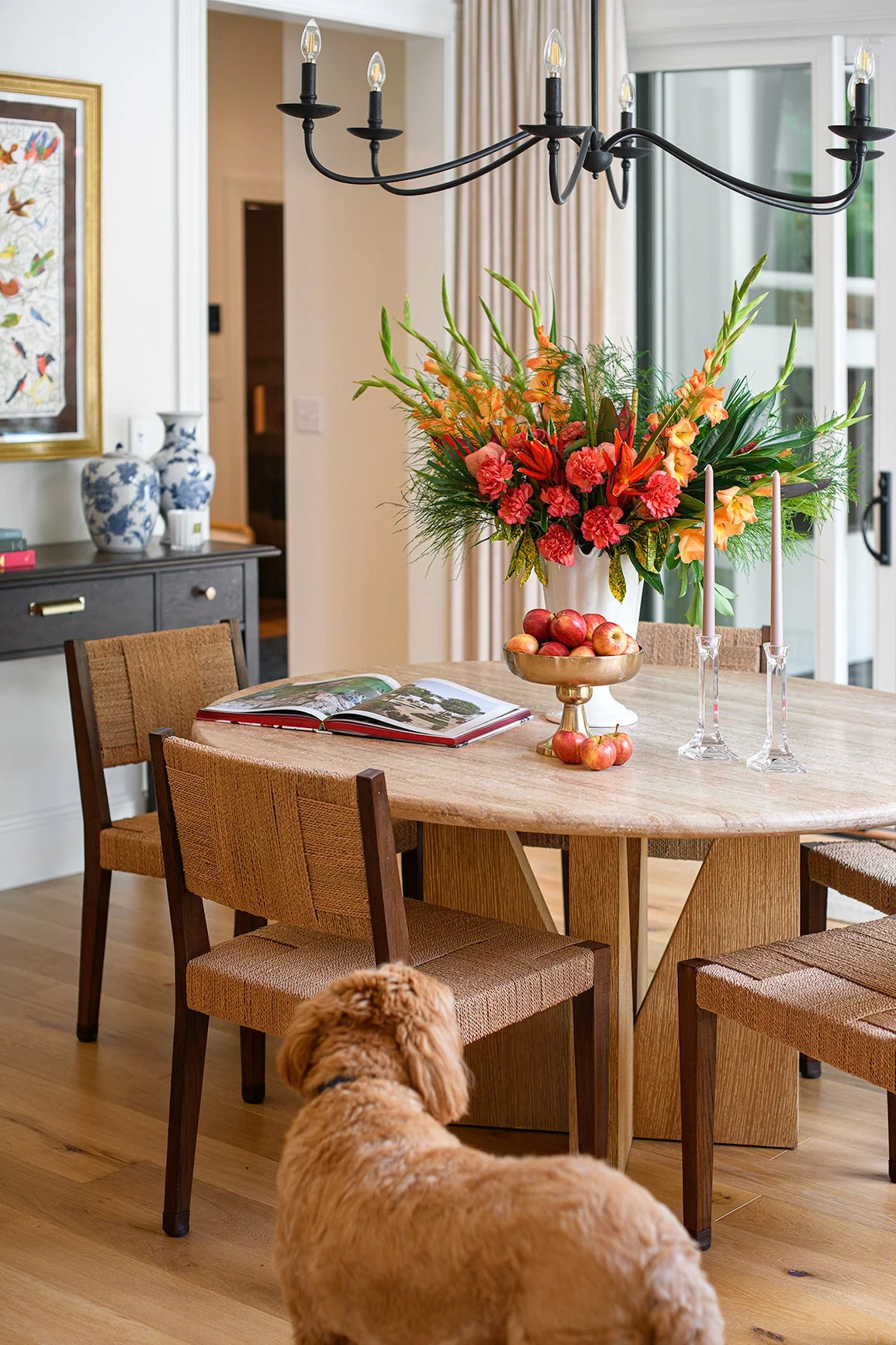 A dining room with a round wooden table, surrounded by chairs with woven seats. There is a large floral arrangement with orange and pink flowers in a white vase on the table, along with a gold bowl filled with small apples, and two pink candles in gl