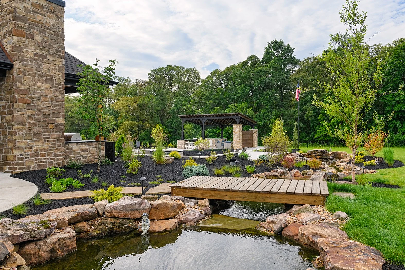 Backyard with a pond and a small wooden bridge, landscaping with plants and rocks, and a covered outdoor seating area with a stone fireplace in the background.