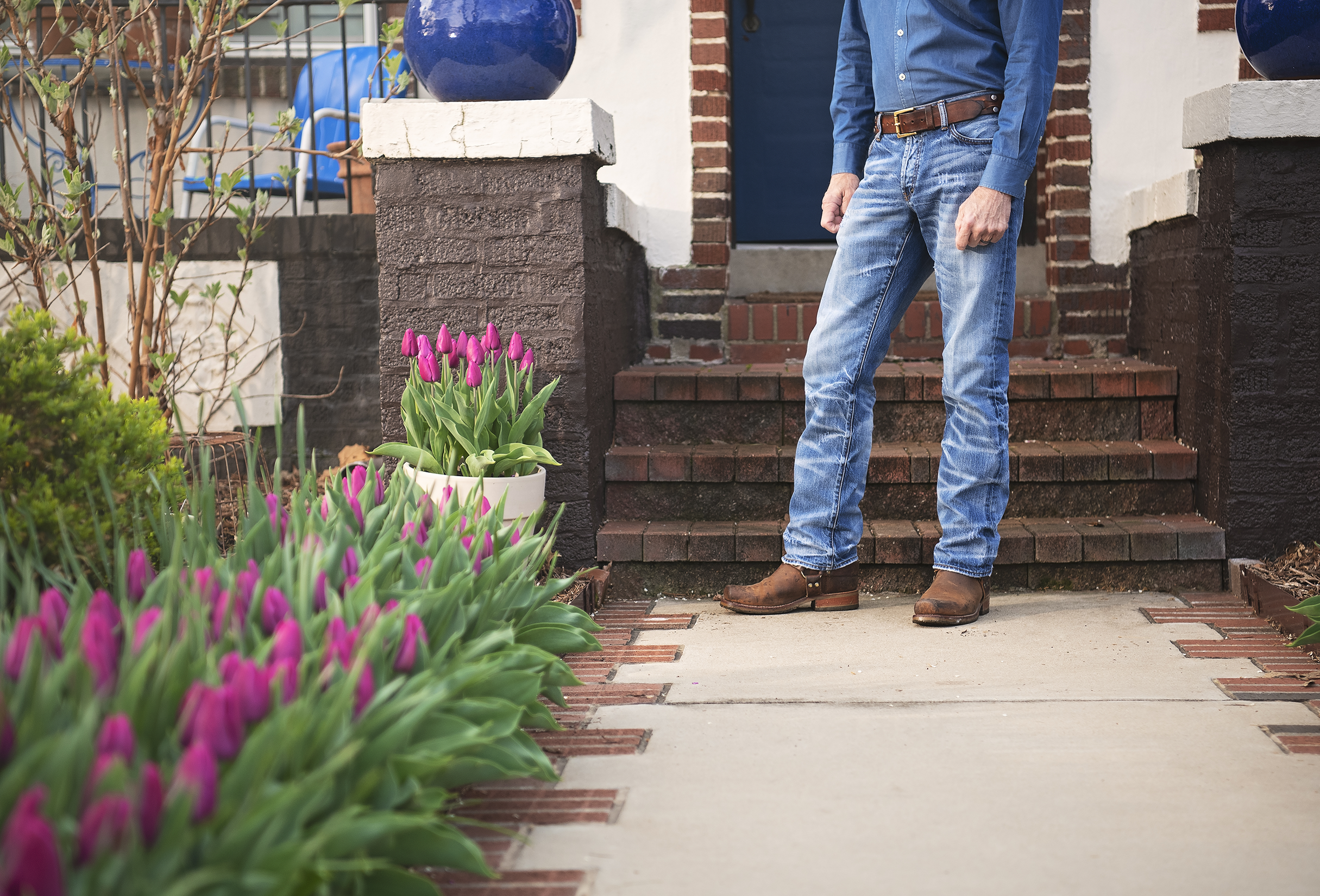 Person standing outside on the front porch wearing a blue shirt, jeans, and brown boots. There are pink tulips in a white pot and a row of tulips along the walkway leading to brick steps in the front yard.