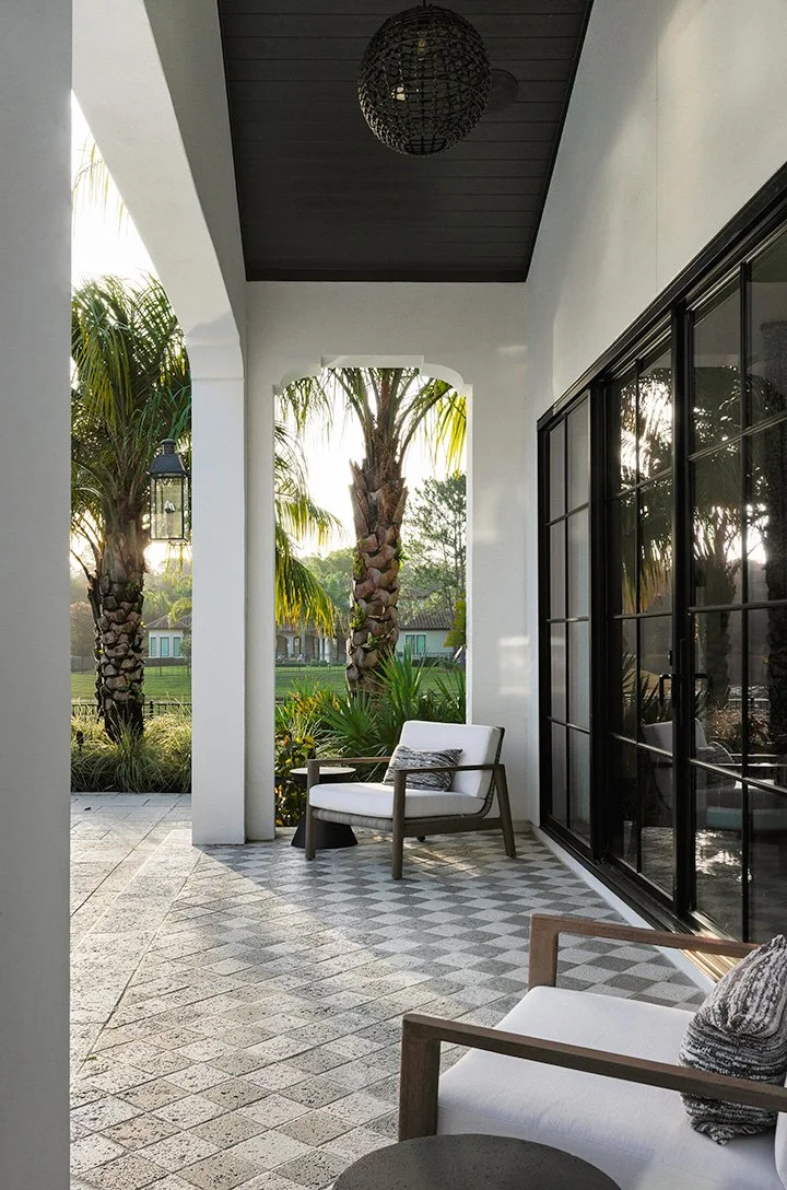 Covered porch with white walls and black ceiling, featuring two white cushioned chairs with wooden frames, a patterned cushion, and a small black side table. Large black-framed glass doors reflect the outdoor greenery, including palm trees, with a gr