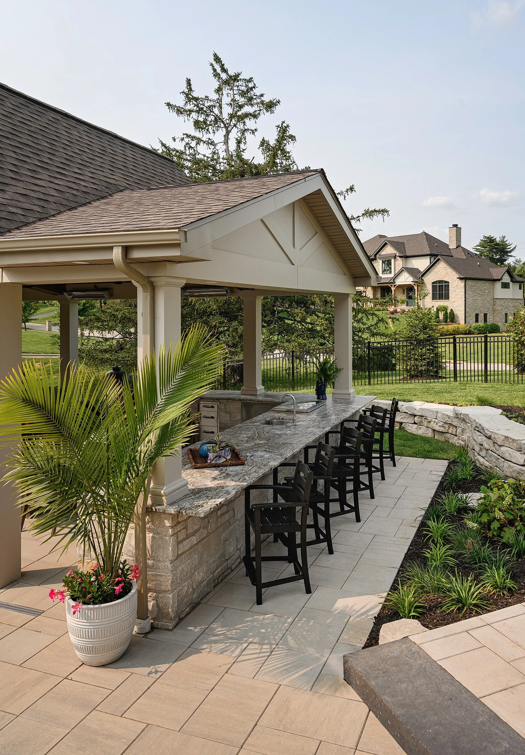 Outdoor kitchen with a stone counter and black chairs under a pavilion, surrounded by landscaped garden and residential neighborhood.