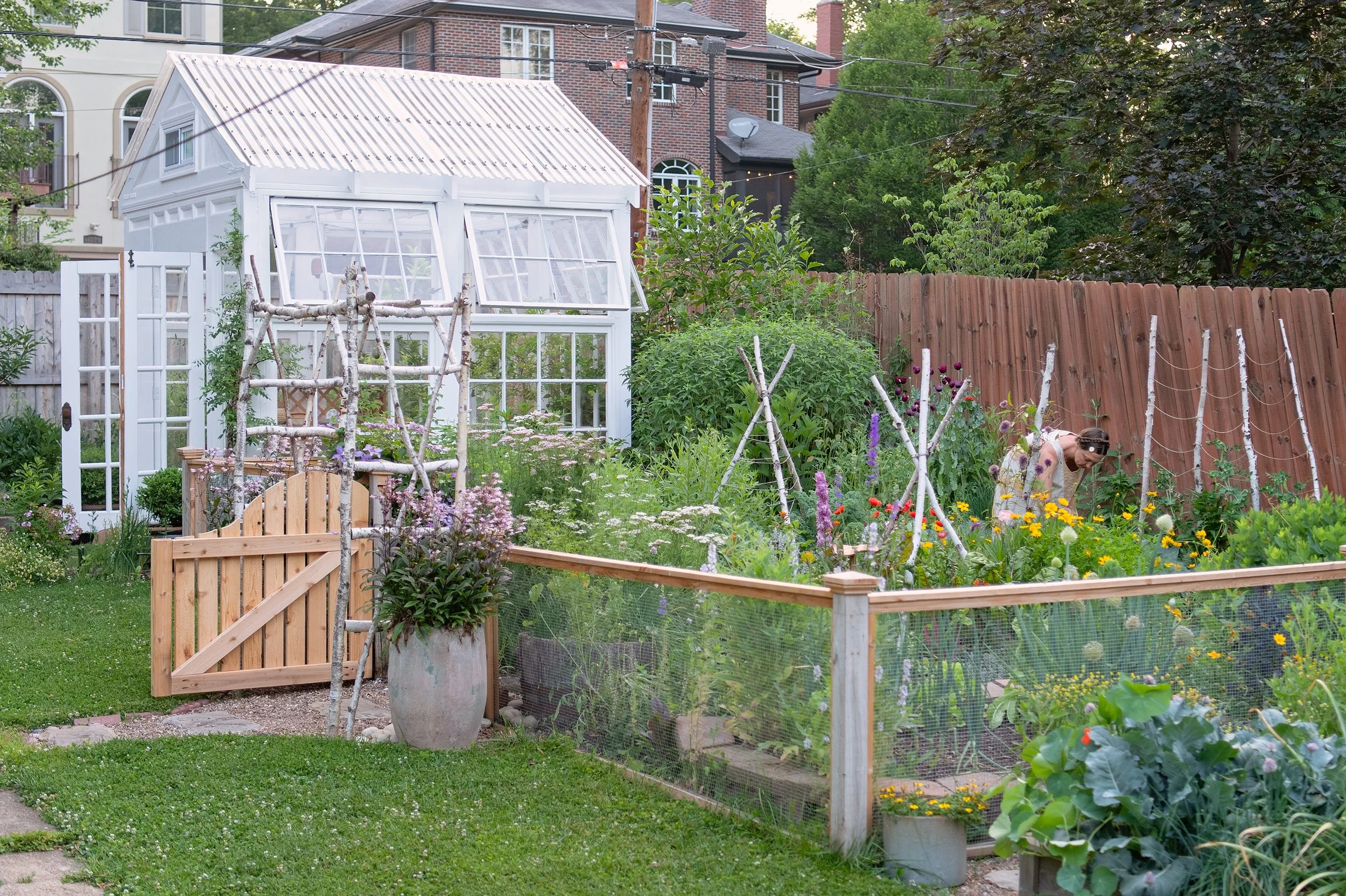 A person tending to a lush vegetable garden enclosed by a wire fence, with a white greenhouse and various flowers and plants, surrounded by a wooden fence and greenery.