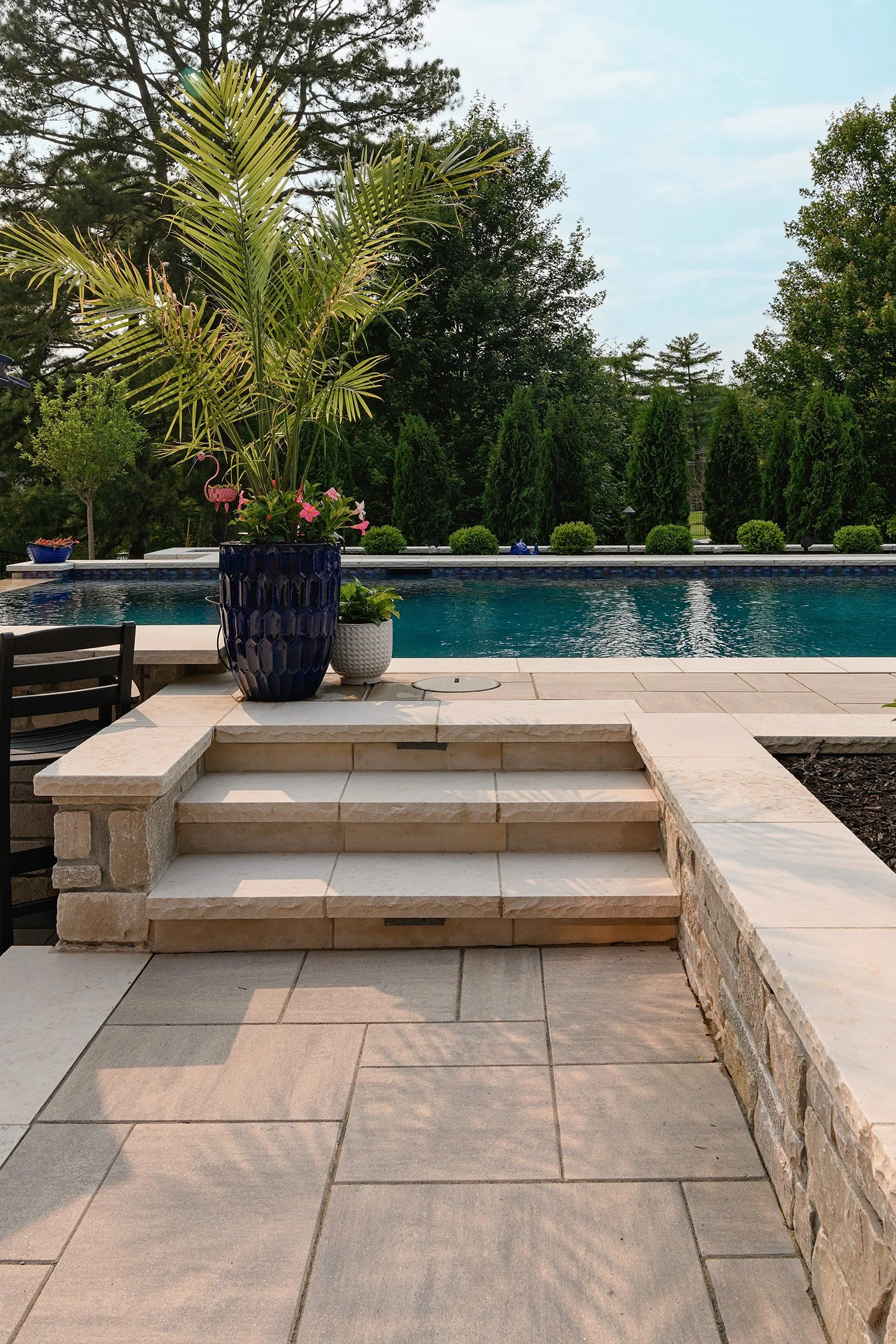 A backyard swimming pool surrounded by a stone patio, with potted plants and greenery in the background.