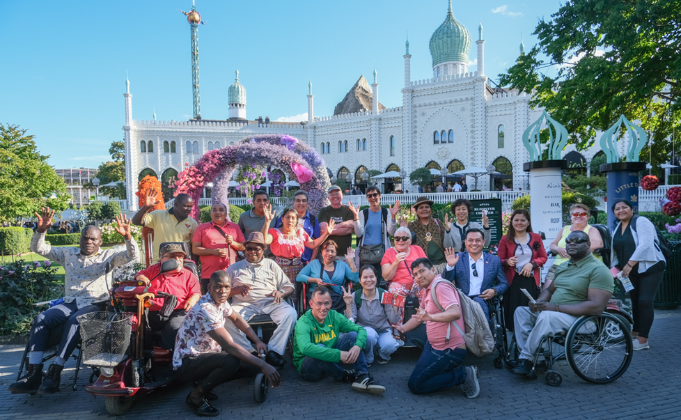 fællesbillede af personer der vinker til kameraet. I baggrunden ses en hvid bygning der ligner et eksotisk palads