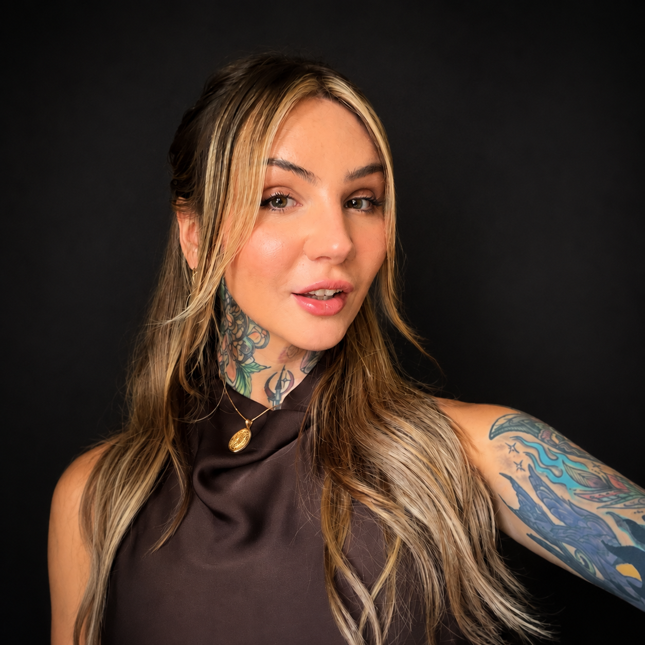 A woman with long, wavy blonde hair and tattoos, wearing a dark satin sleeveless top and a gold pendant necklace, is taking a selfie against a black background.