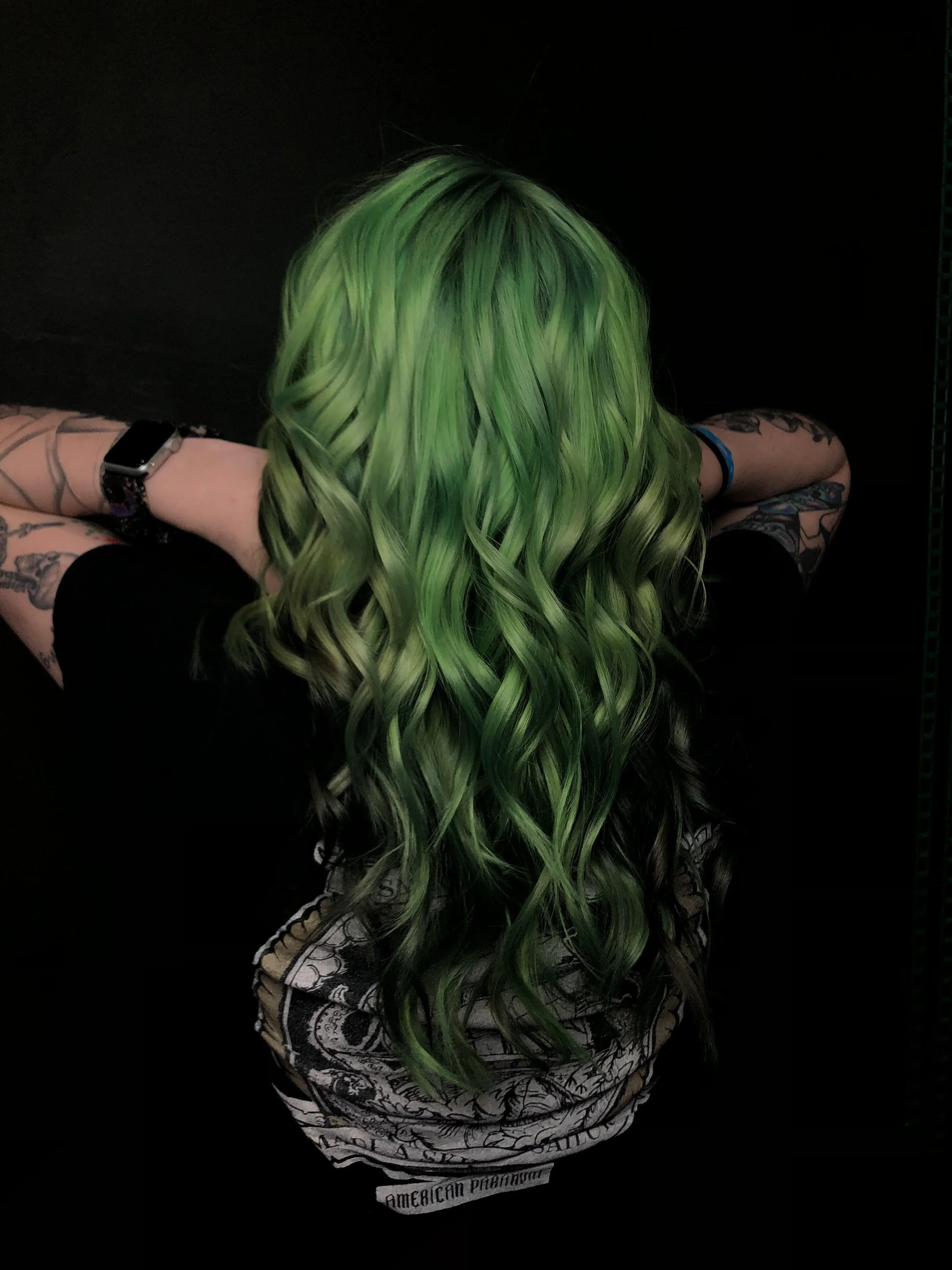 Person with long, wavy green hair and tattoos, wearing a black shirt and a smartwatch, leaning forward with arms resting on a surface against a black background.
