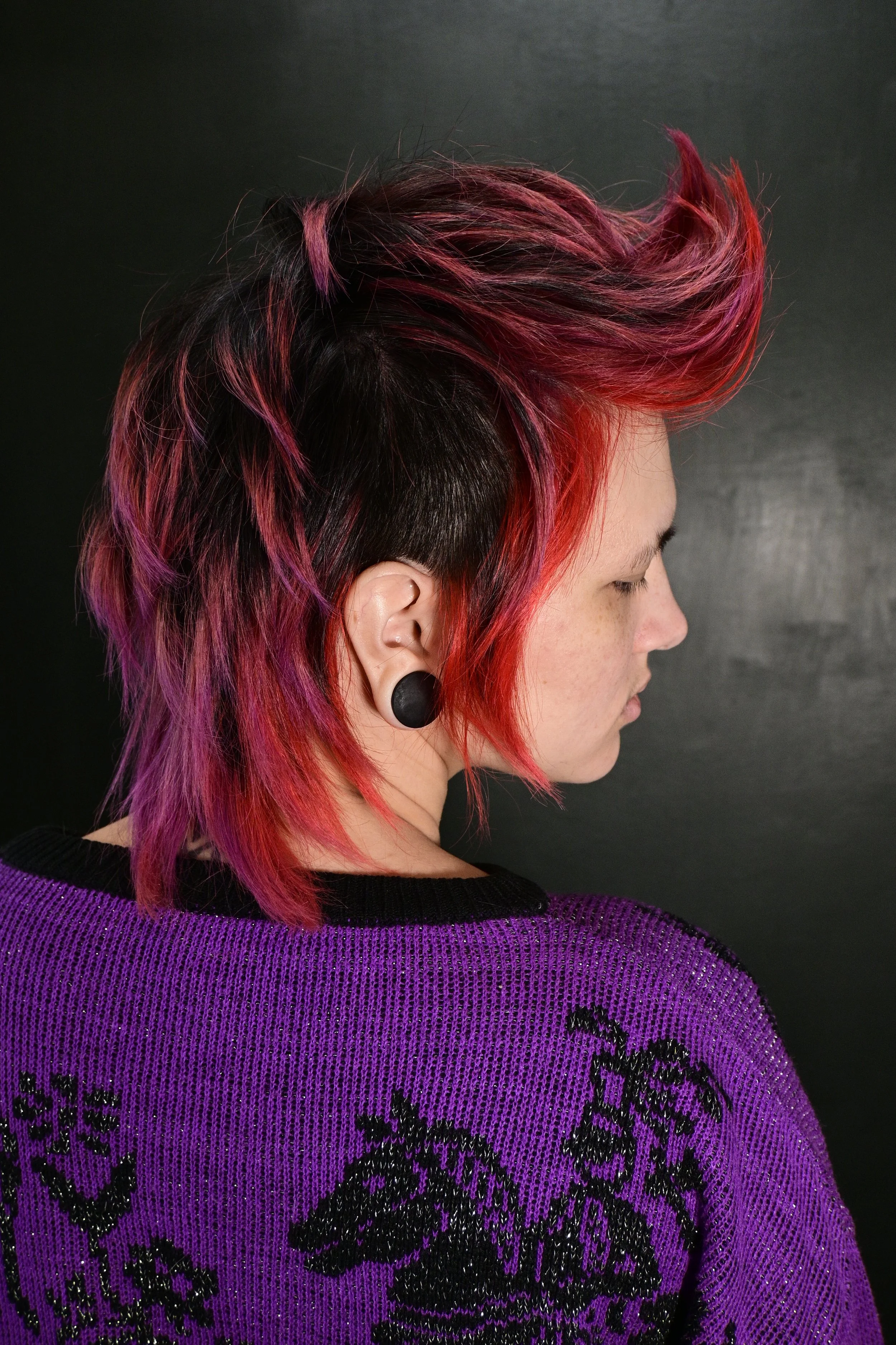 Side profile of a woman with short, spiky hair dyed in shades of pink and purple, wearing a purple sweater with black embroidered floral patterns, against a black background.