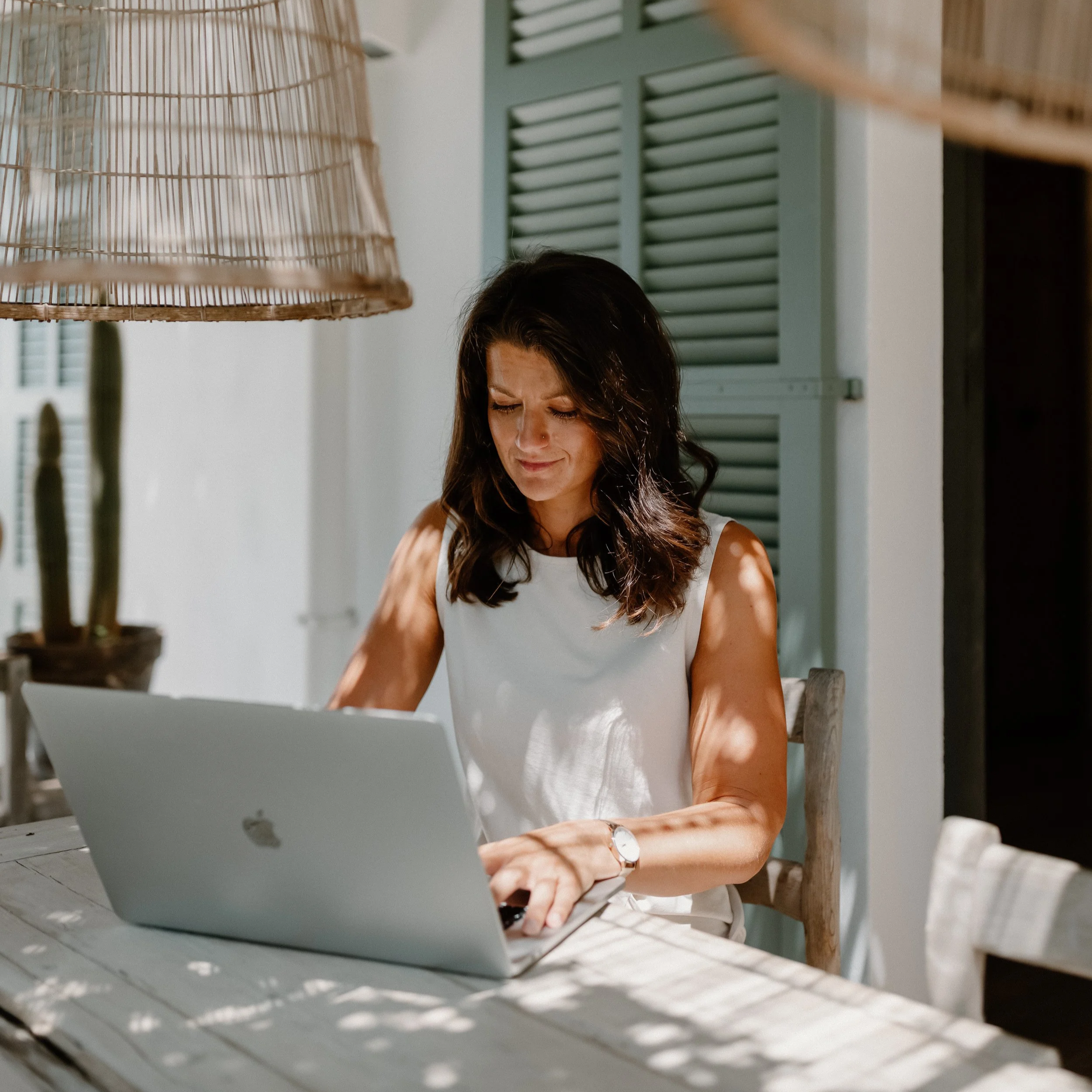 Woman working on a laptop at a rustic table on a sunny patio with rattan lamps and green shutters.