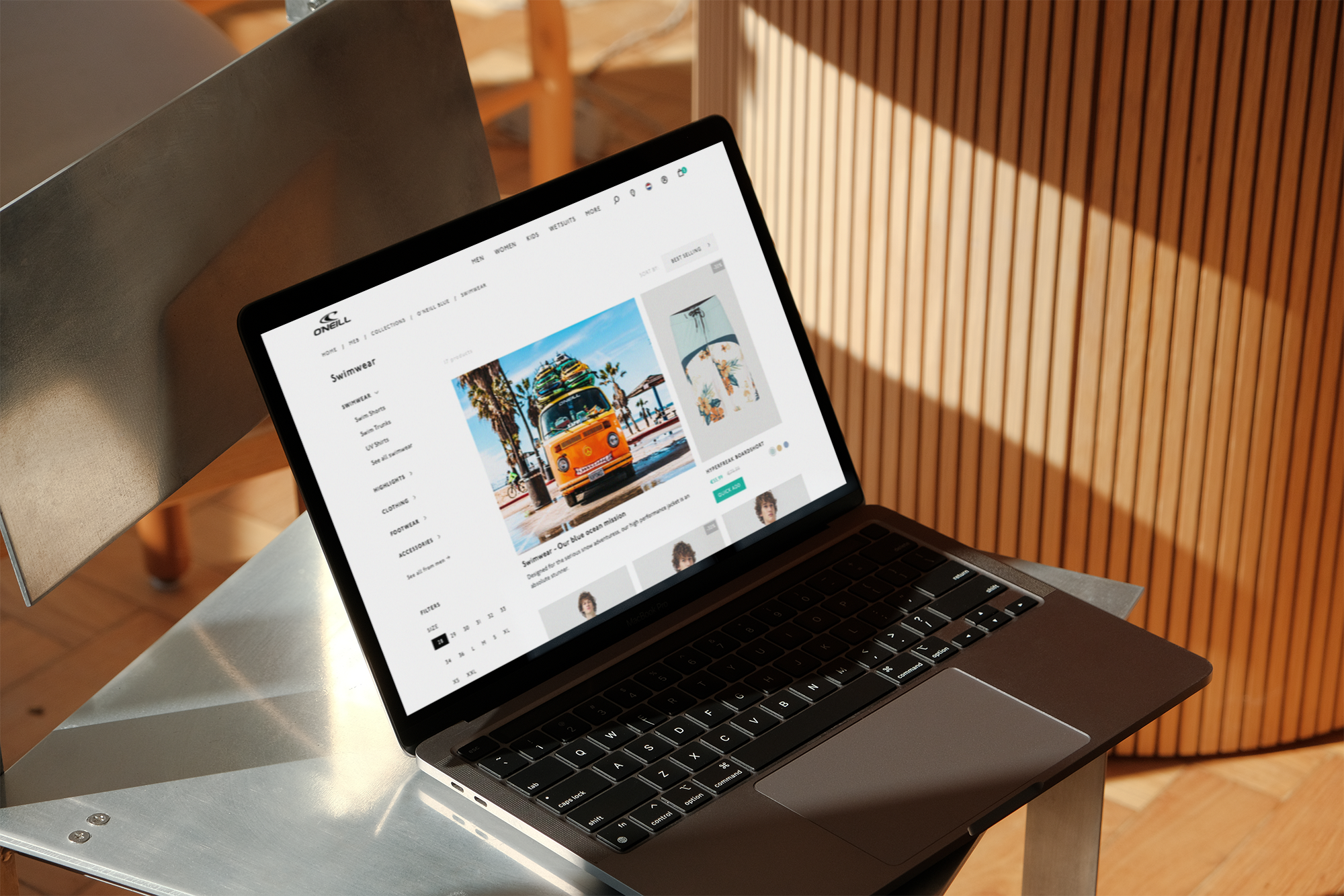 Laptop on a small metal table displaying an online store for swimwear. The page shows a picture of a yellow van with surfboards on top, palm trees, and various swimwear items.