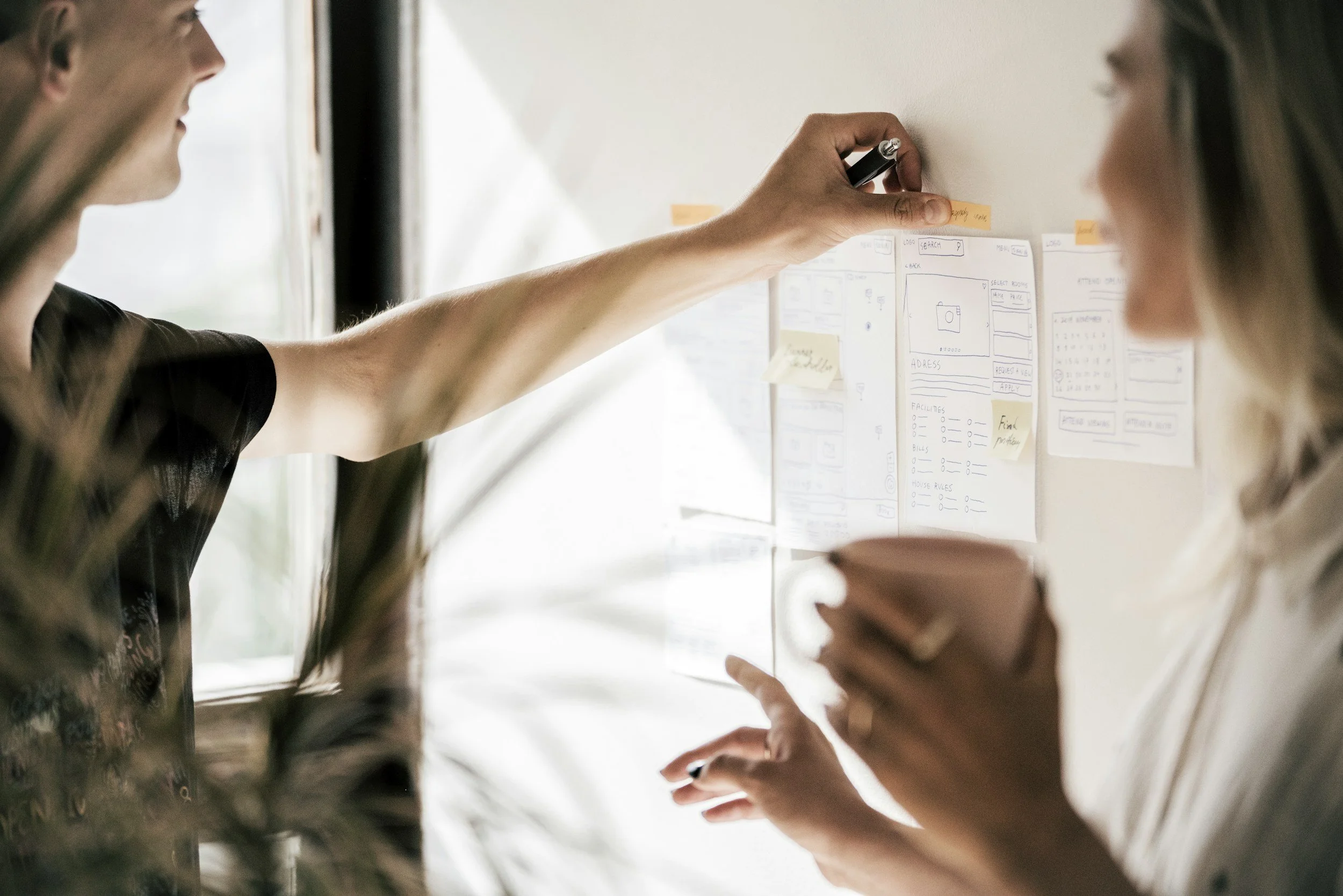 A person writing on sticky notes on a whiteboard with another person holding a phone, in a collaborative work setting.