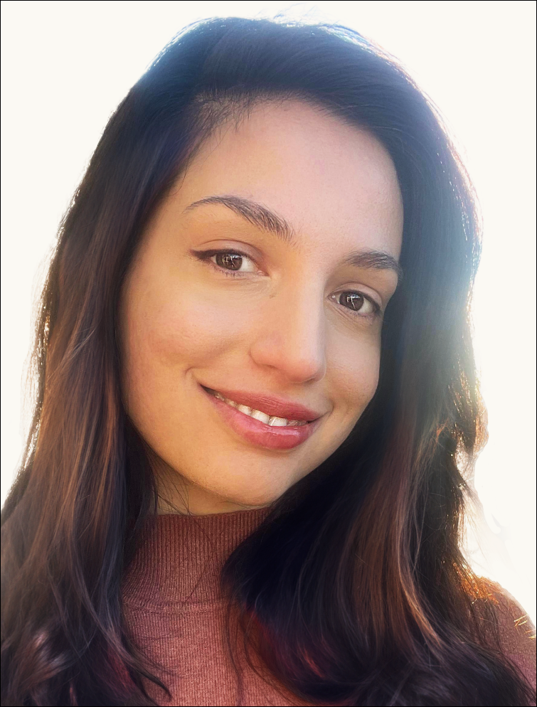Close-up of a young woman with dark brown hair, smiling at the camera, wearing a brown top, with sunlight in the background.