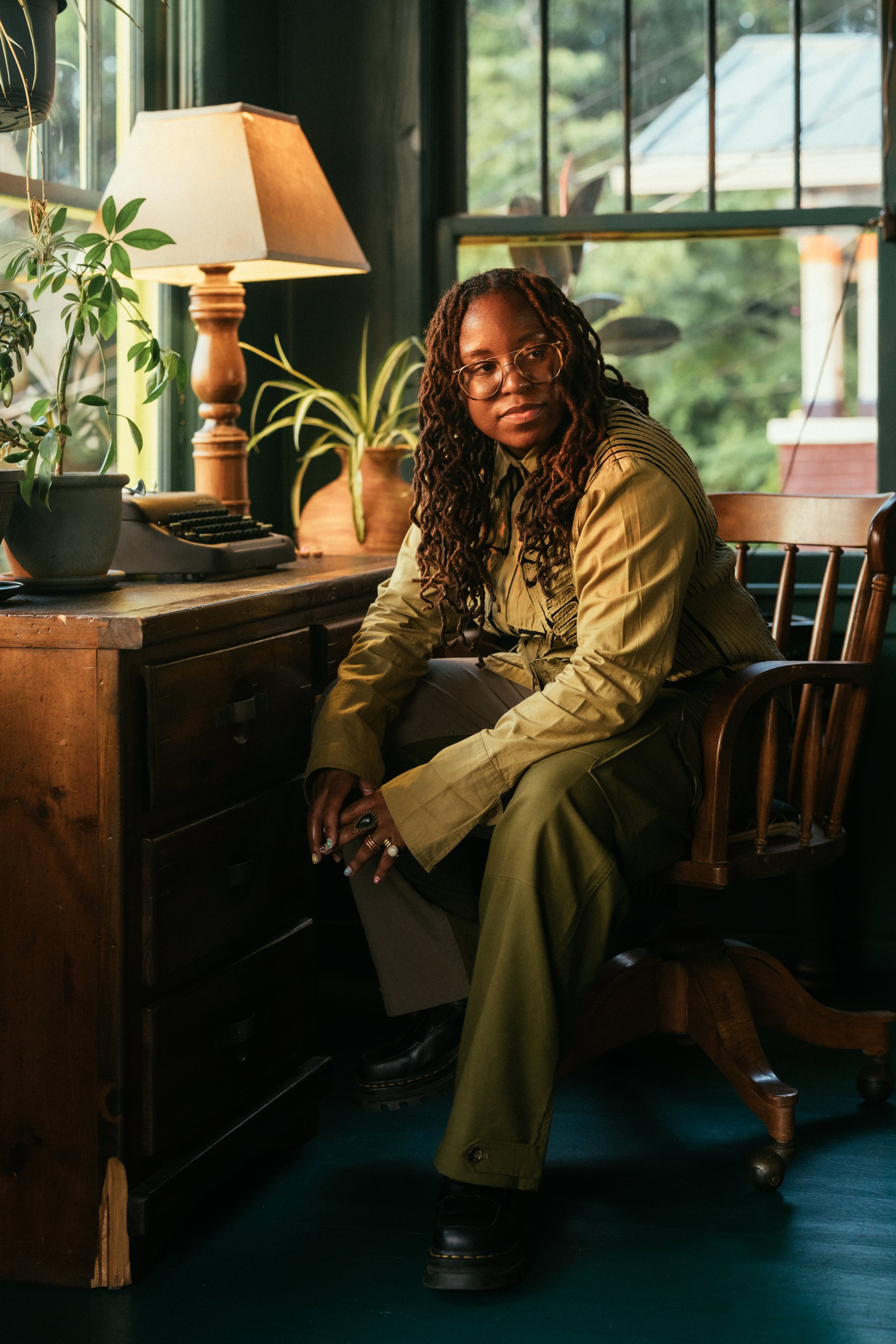 Portrait of a non-binary person with long locs sitting in a wooden chair and antique desk, warmly lit by a table lamp in a dark green room filled with plants. They wear an olive-green outfit and boots, looking out of frame in a calm, reflective pose.