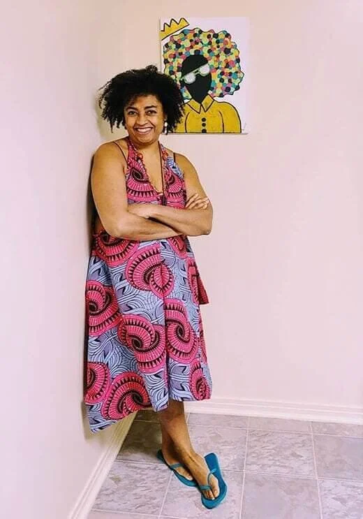 A Black woman with curly black hair smiling wearing a pink and purple patterned dress against a light pink wall.