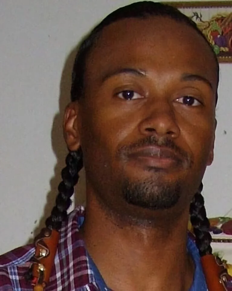 A Black, Indigenous man wearing two braids against a dim background in a burgundy , plaid, collared shirt.