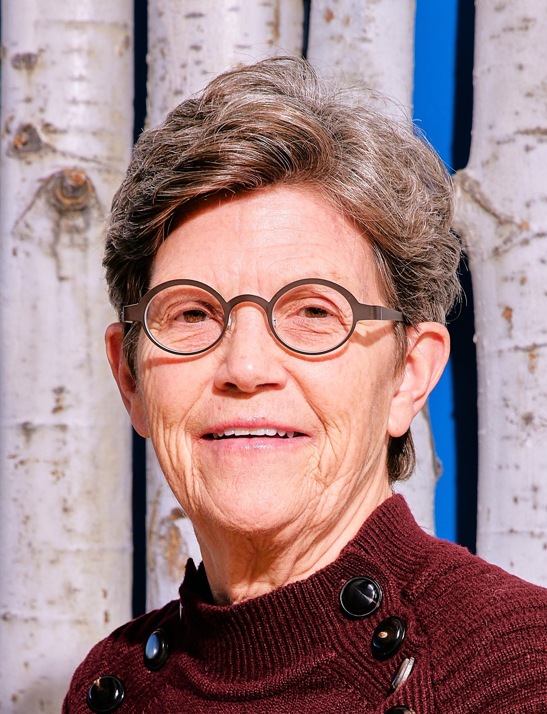 A smiling white woman with brown and gray hair, smiling wearing oval glasses and a knitted, burgundy sweater.