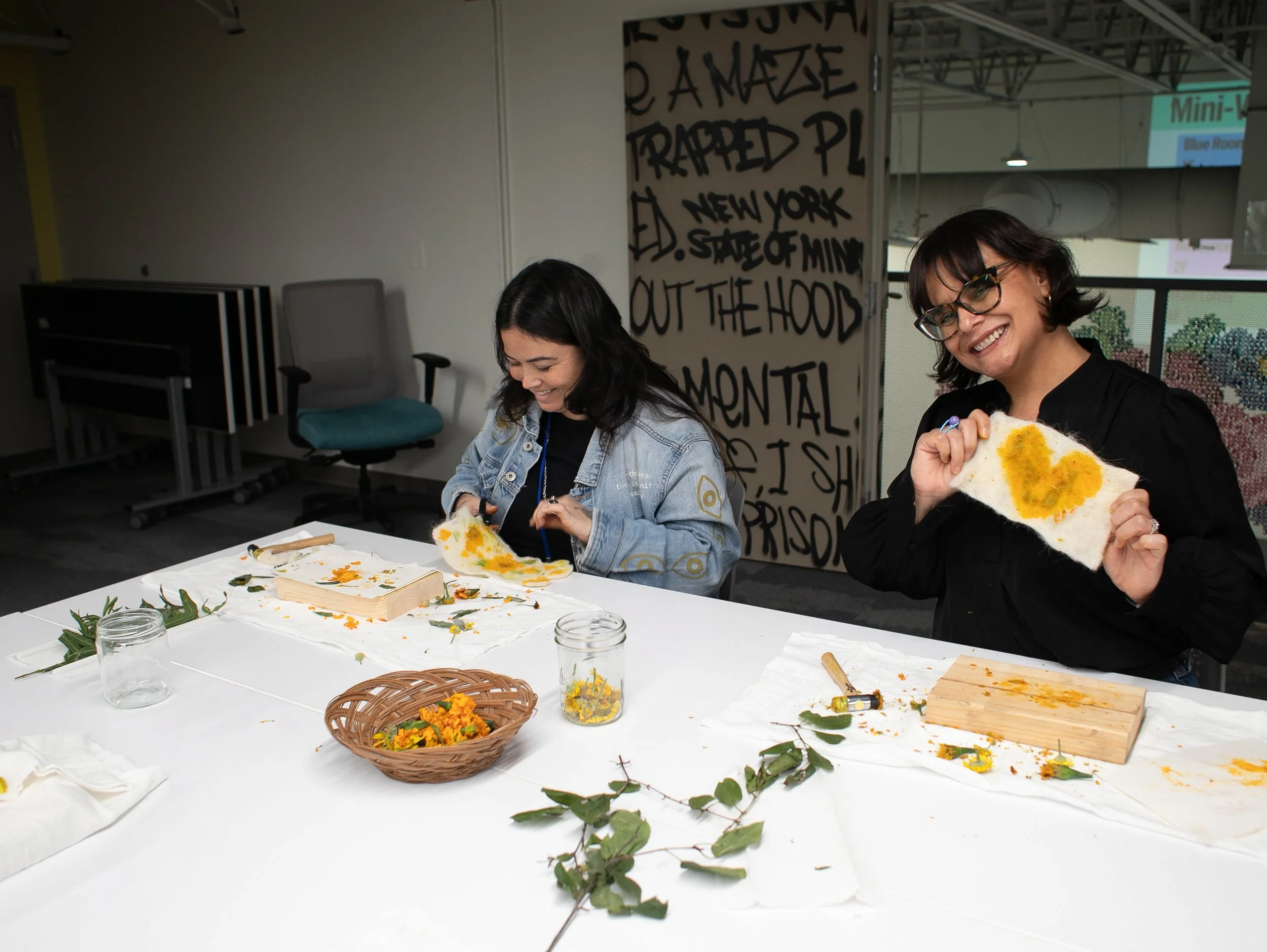 Two women with dark hair, one with glasses, one with a denim jacket; sitting at a white table creating art canvases with dried flowers, leaves, and other craft supplies.