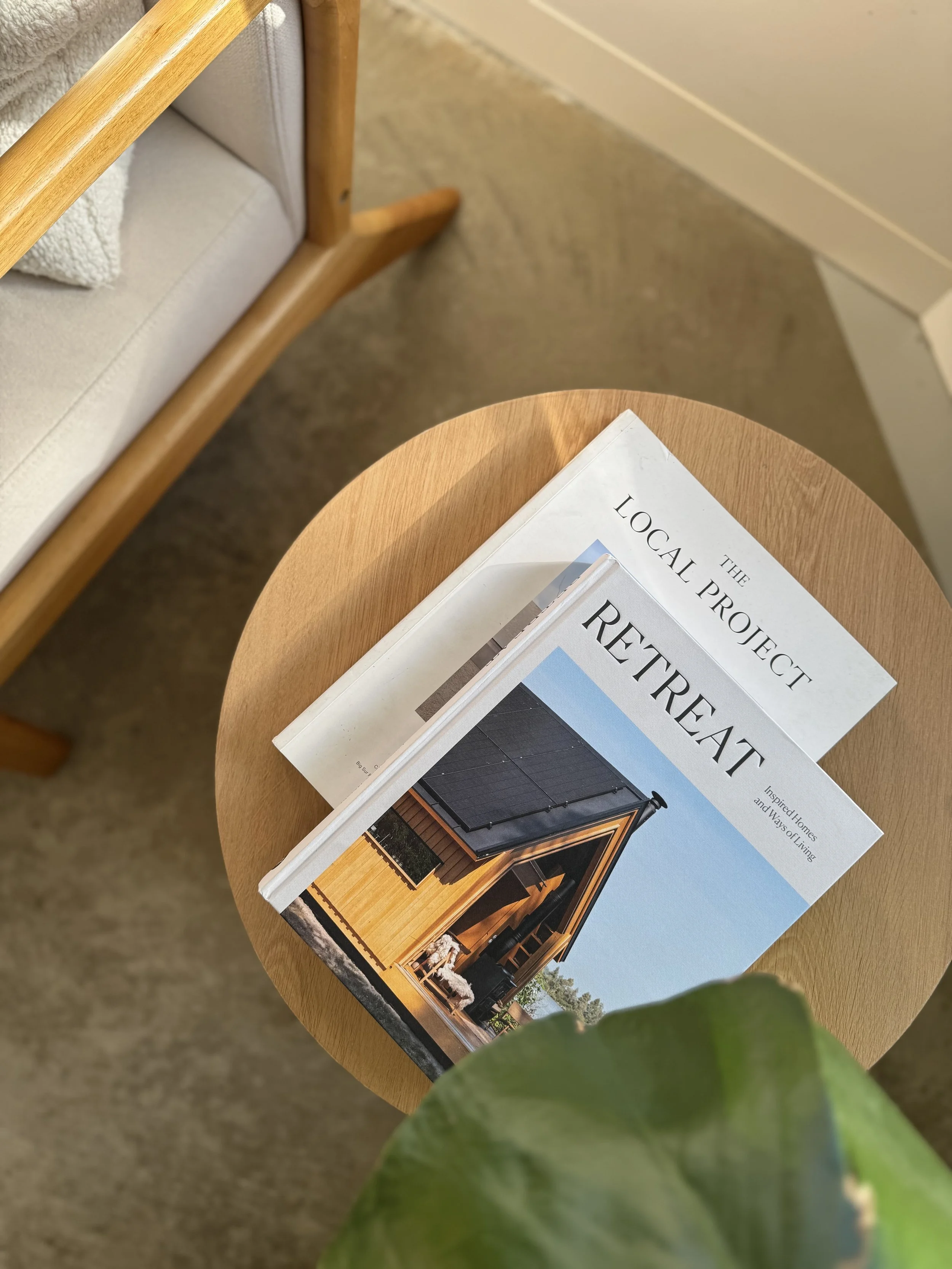 A wooden side table holding four interior design and architecture magazines, with a beige armchair and a green leaf in the foreground and a beige carpeted floor.