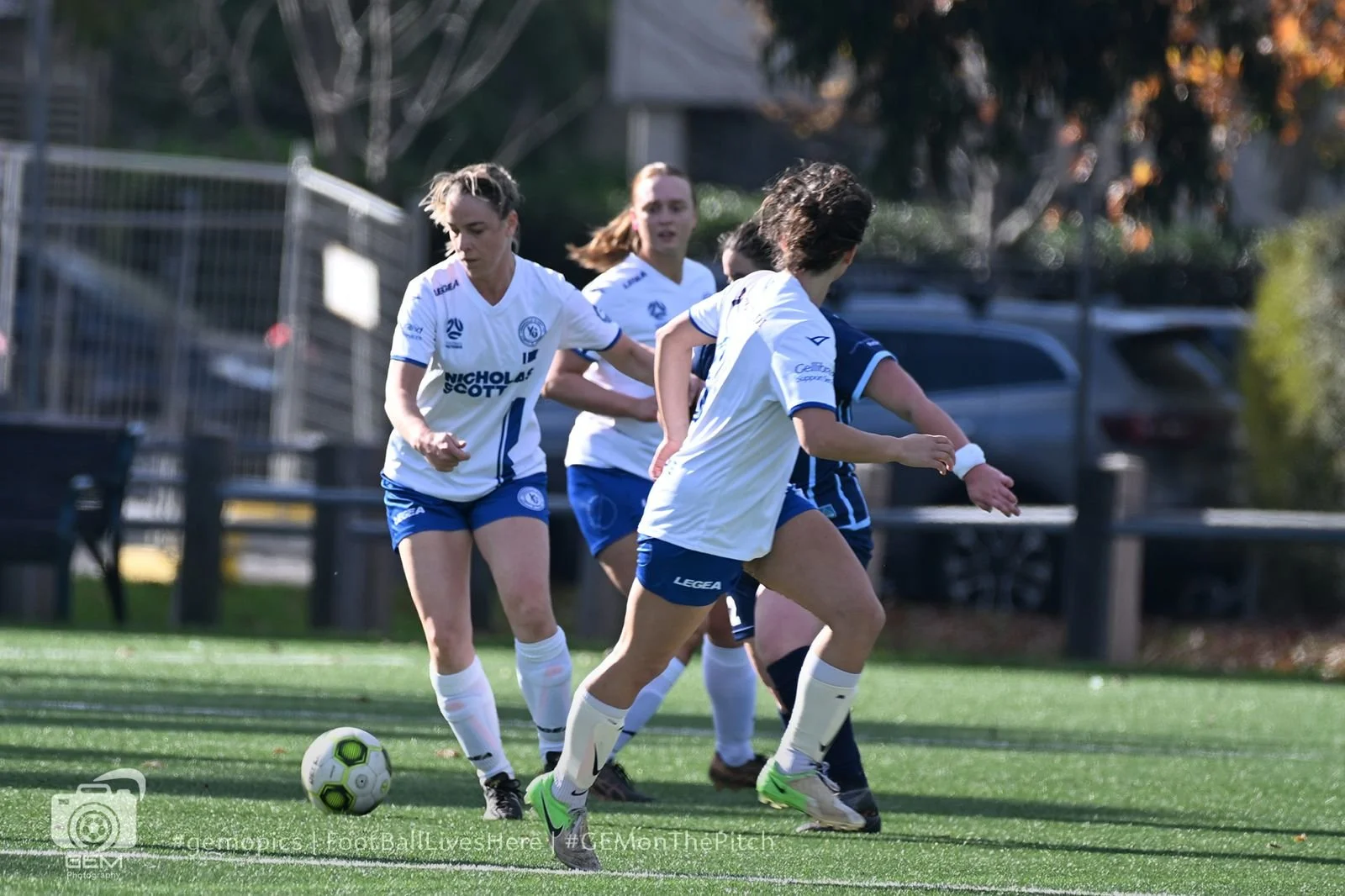 Four female soccer players, wearing white and blue uniforms, battle for ball on the field during a match on a sunny day.