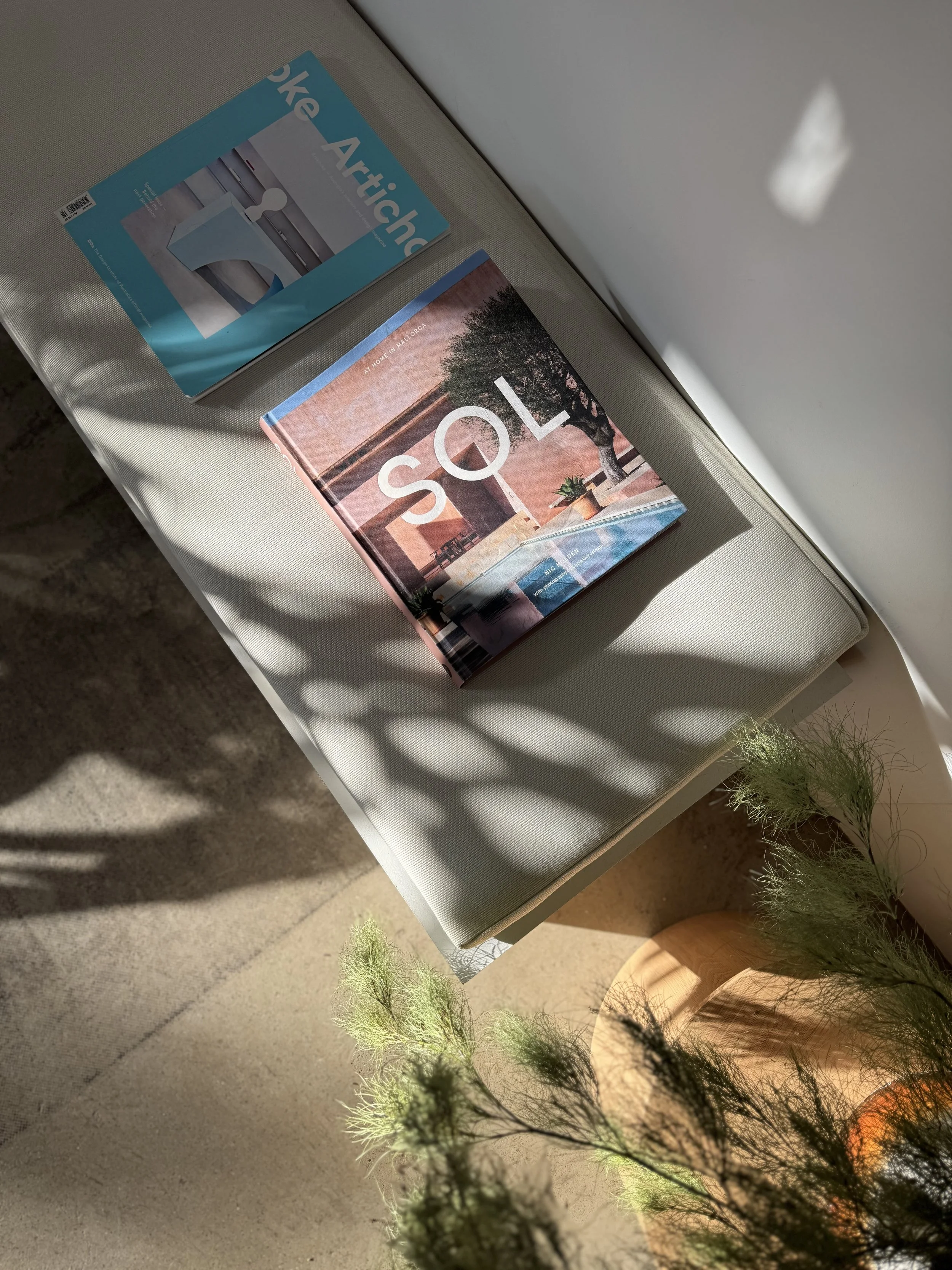 A magazine titled 'SQL' and another magazine titled 'Make Architectural' are placed on a light-colored cushioned bench near a window with sunlight casting shadows. A potted plant with delicate green foliage is nearby.